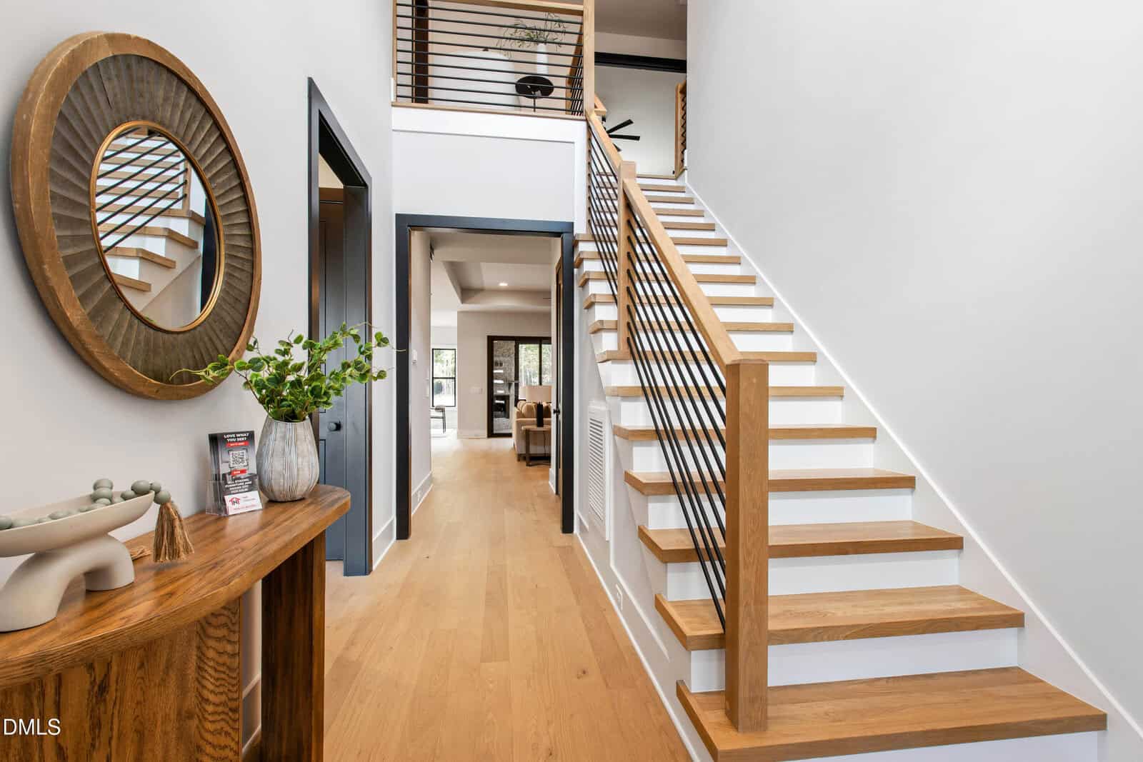 Modern entryway with light wood floors, a wooden staircase with black railings, a round mirror above a console table with decor, and an open view into a bright living area.