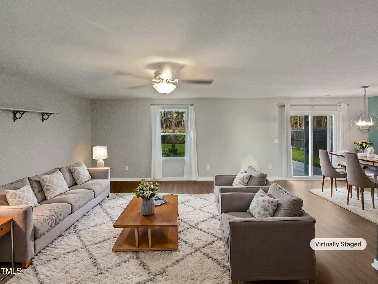 A virtually staged living room features two gray sofas, a wooden coffee table on a white rug, and a ceiling fan. A dining area is visible in the background with a table and chairs. Large windows offer a view of the backyard.