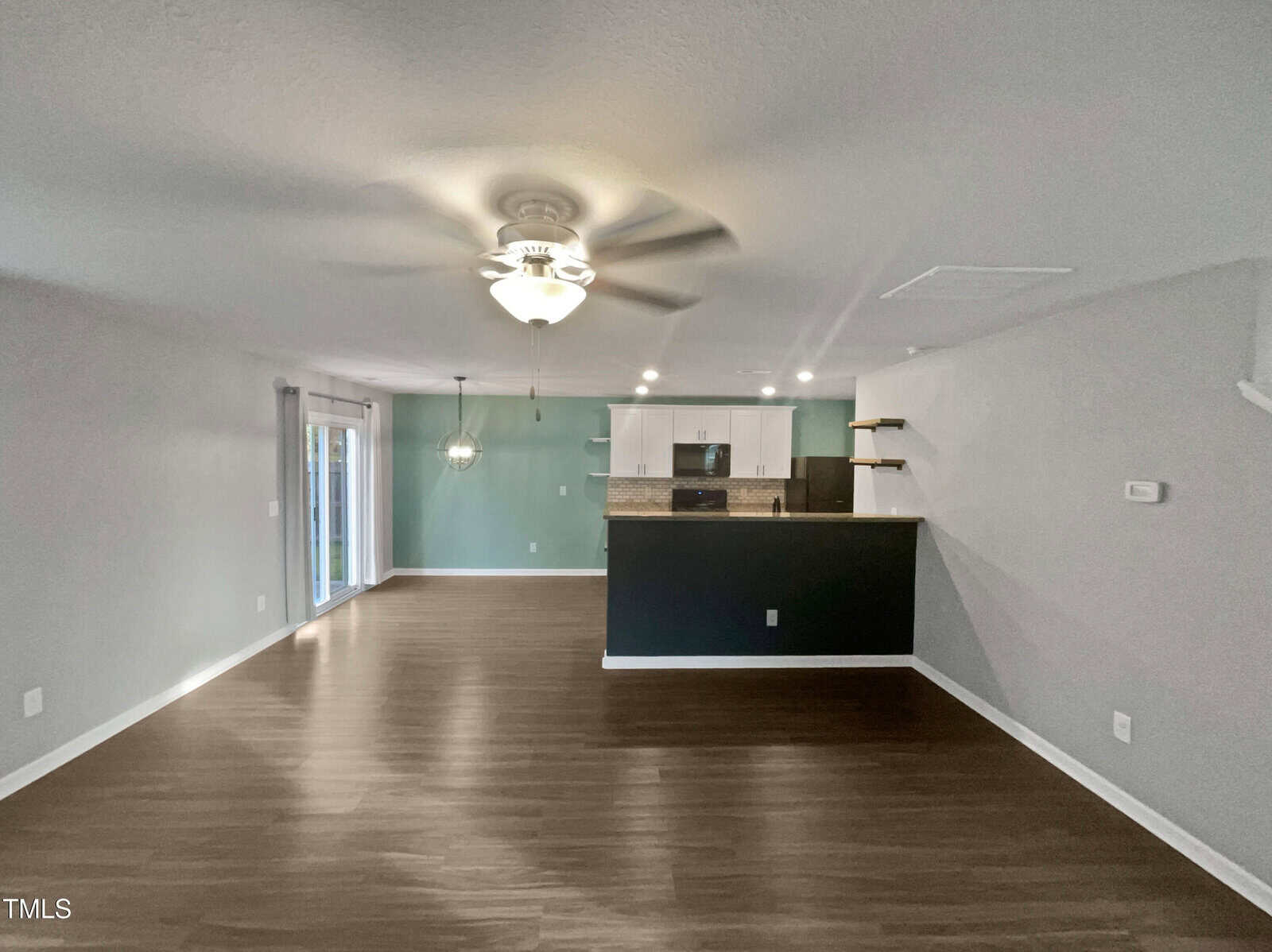 Open-plan living area with wood flooring, featuring a ceiling fan and a hanging light fixture. The room leads to a kitchen with a breakfast bar and stainless steel appliances. Sliding doors on the left open to a patio area. Walls are painted light gray.
