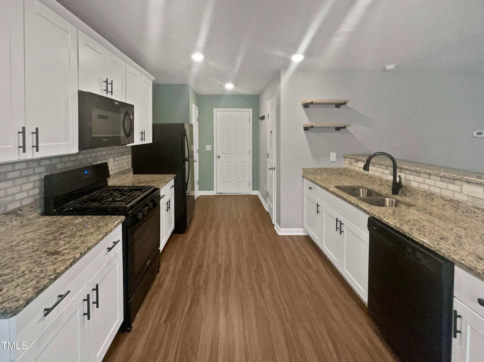 A modern kitchen with white cabinets, granite countertops, and wooden floors. Includes a black stove, refrigerator, and dishwasher. The walls are light gray, featuring open shelves above the sink, and a doorway near the kitchen's end.