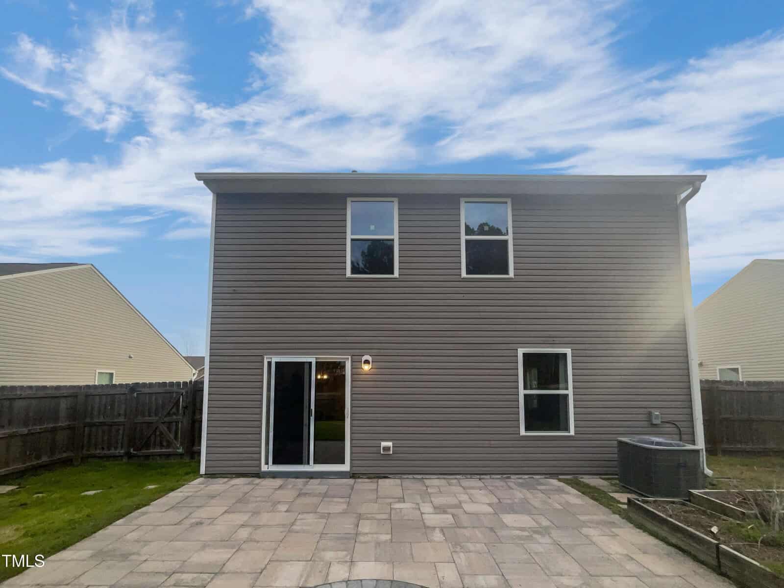 The image shows the back view of a two-story house with brown siding. There are two windows on the upper floor and a sliding glass door on the ground level, leading to a stone patio. The yard is fenced, and the sky is clear with some clouds.