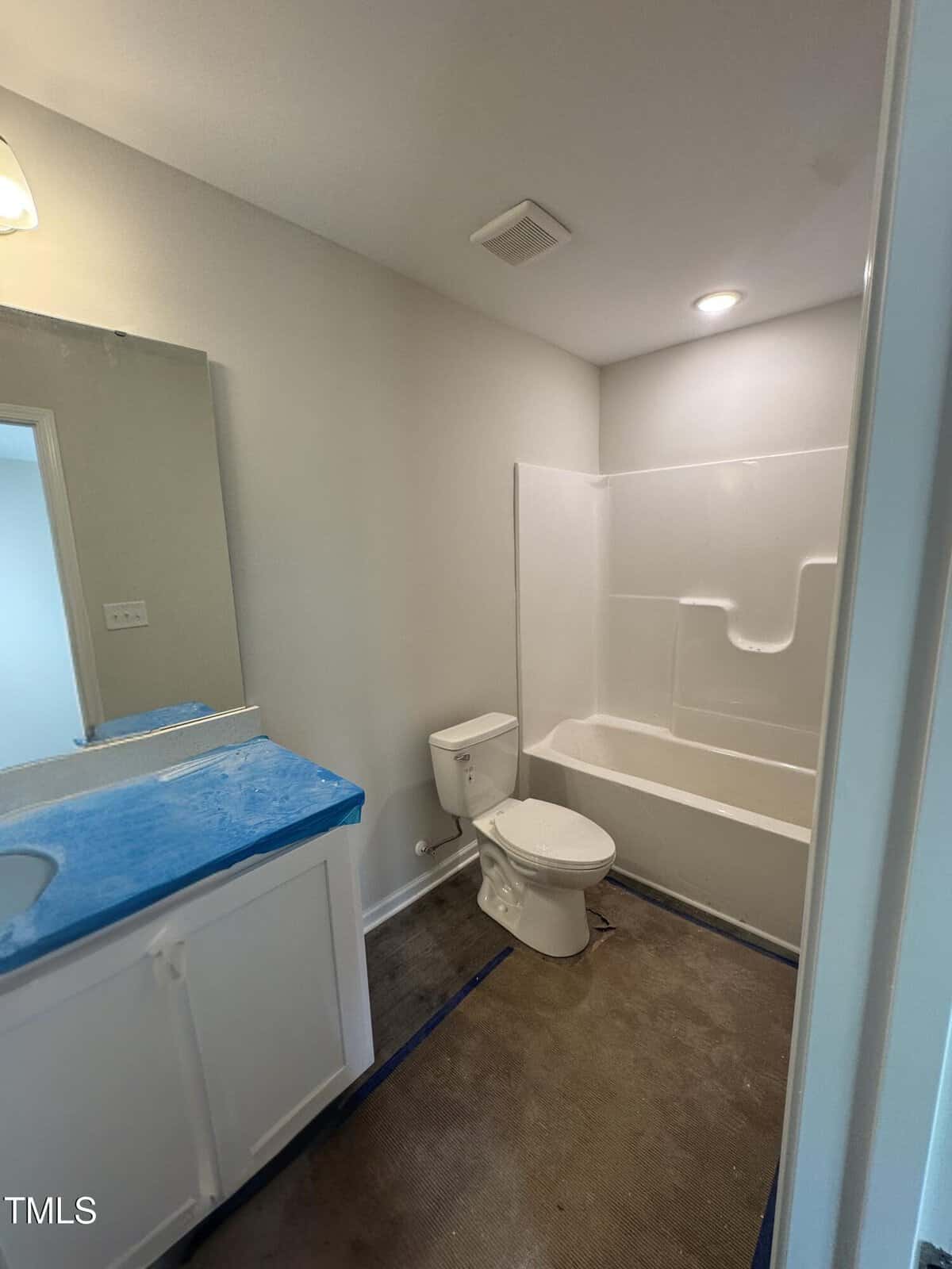A bathroom with a white bathtub and shower combination, a toilet, and a vanity with a blue protective cover. The floor appears unfinished and there is a wall mirror above the vanity.
