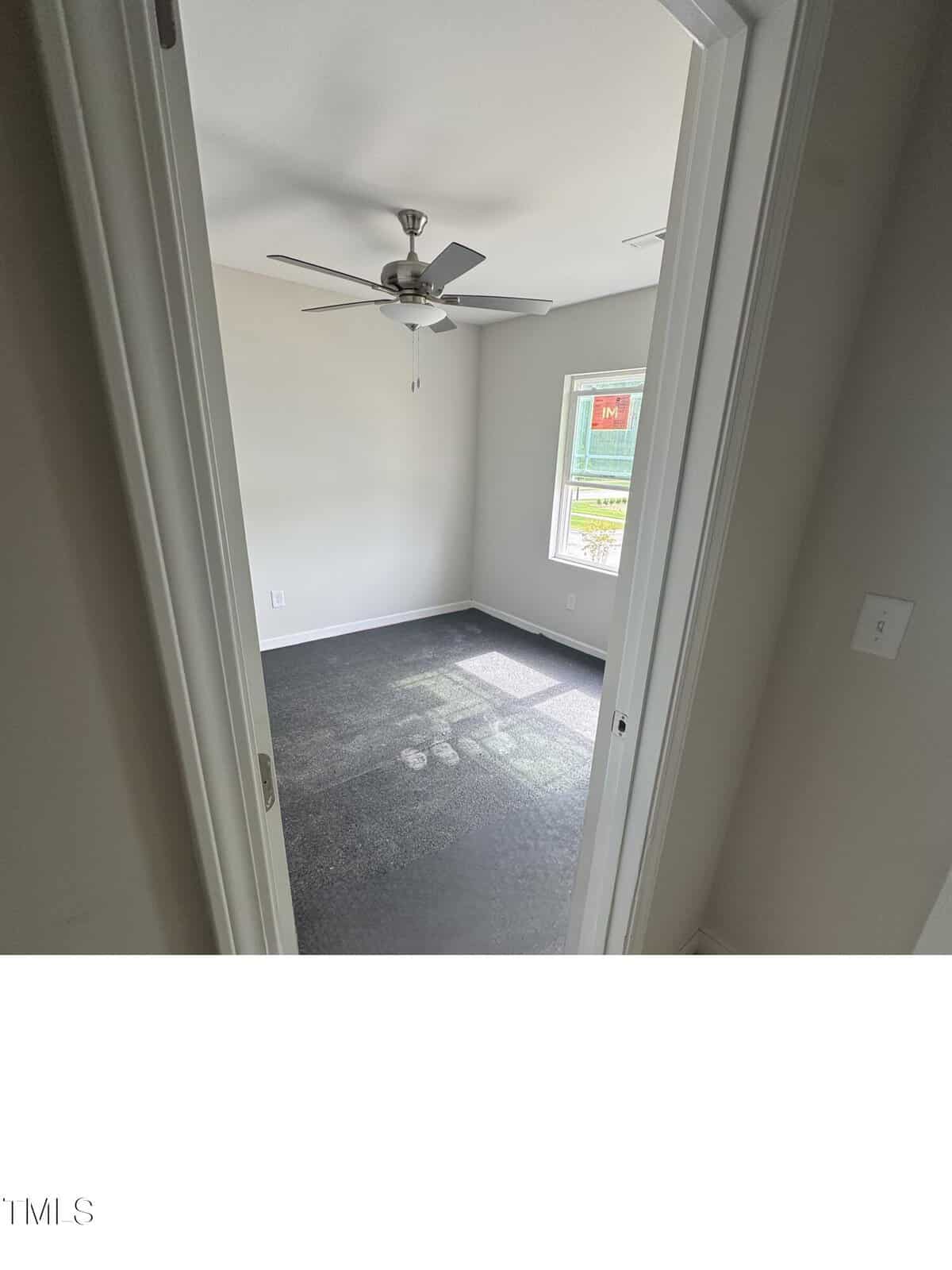 A small empty room with gray carpet, white walls, a ceiling fan, and a window letting in natural light. The photo is taken from the doorway, showing part of the adjacent wall and light switches.