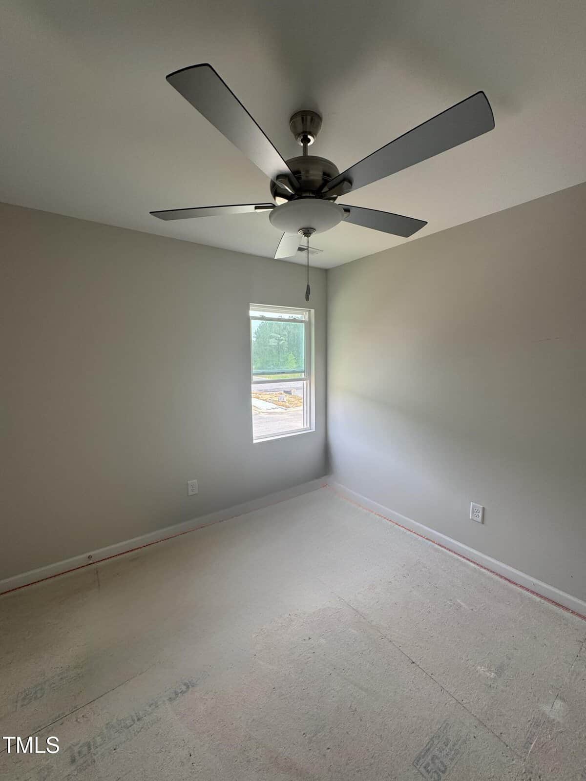 An empty room with light gray walls, a single window, a ceiling fan with four blades, and an unfinished floor. Electrical outlets are visible on two walls. Natural light enters through the window.