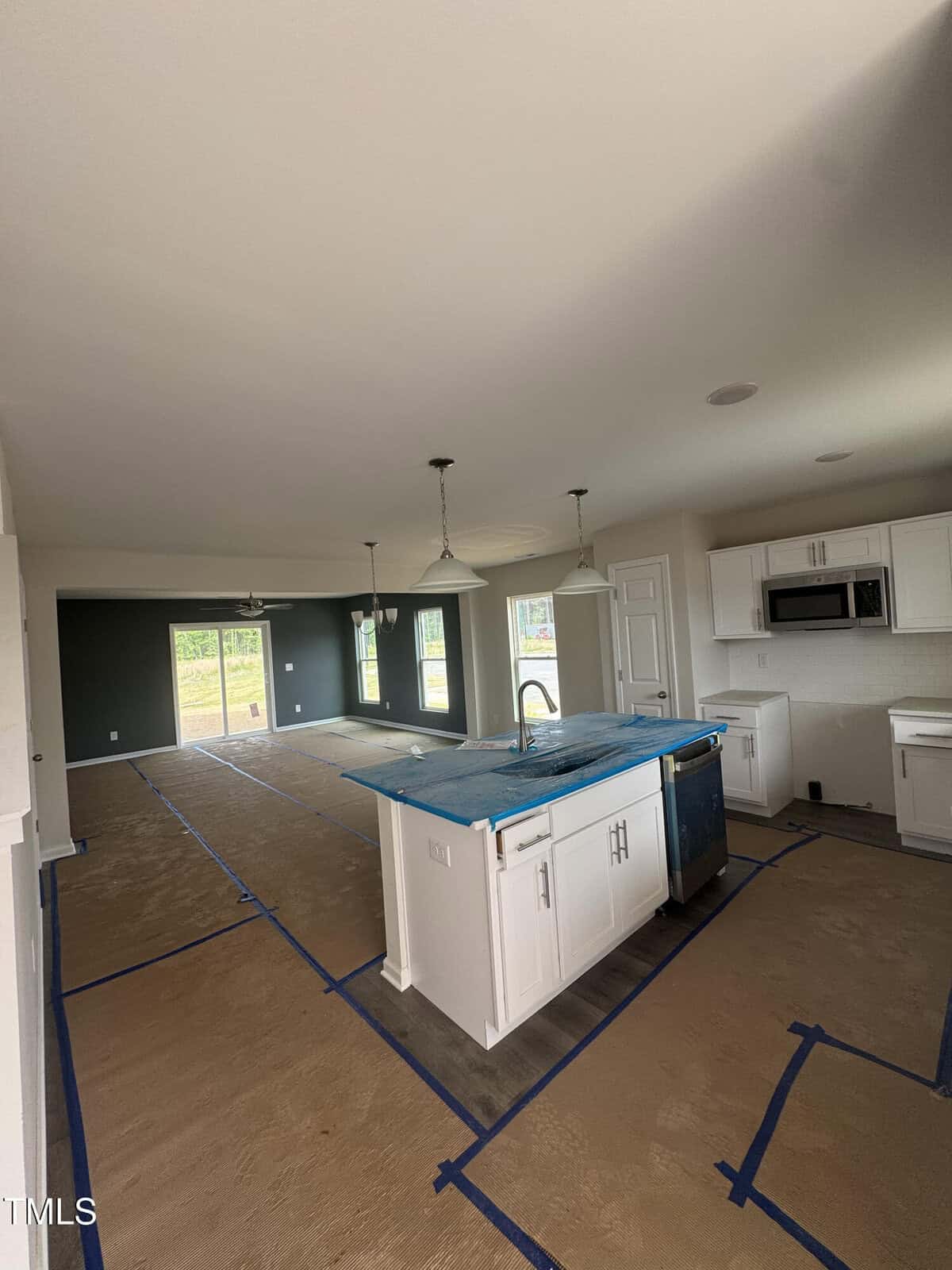 A modern kitchen with white cabinets, a center island, and appliances. The floor is covered with protective brown paper and blue tape. The kitchen opens to a living area with large windows and a glass door leading outside.