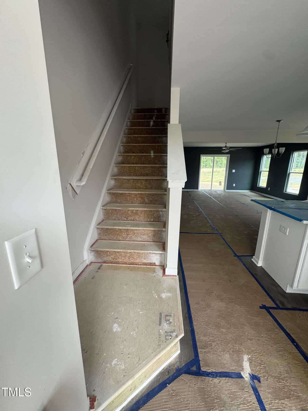 A view of an unfinished staircase with no carpet, leading to an upper floor. The area beside the stairs and the adjacent open living space have protective coverings on the floor. White walls and multiple windows are visible.