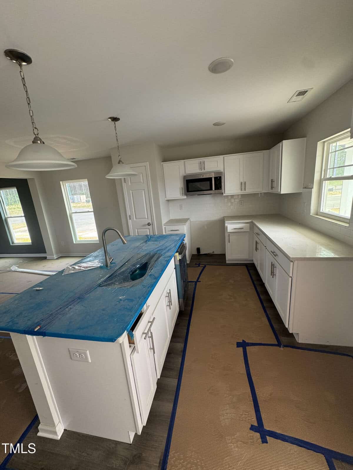 A modern kitchen under construction with white cabinets, a center island covered in blue protective plastic, and cardboard with blue tape on the floor. The room is well-lit with natural light from multiple windows.
