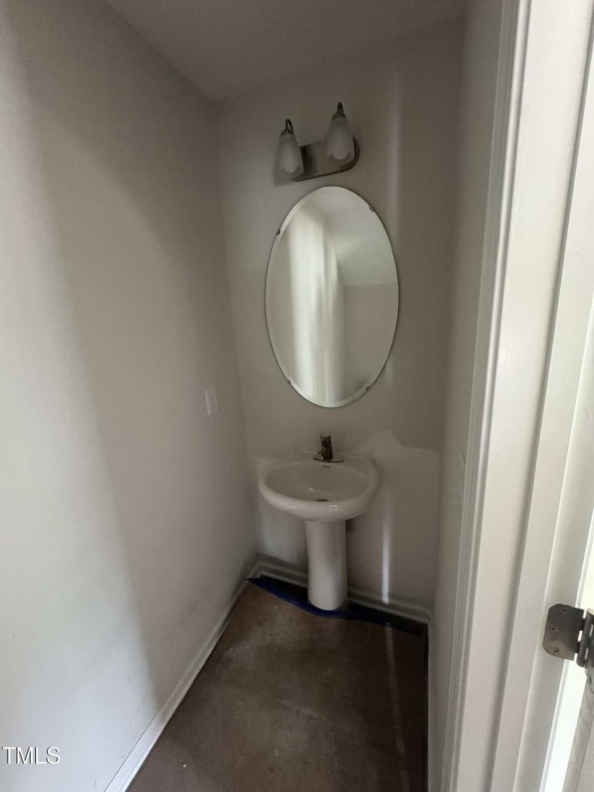 A small, empty bathroom with white walls, a pedestal sink, an oval mirror, and a two-bulb light fixture above. The floor has no visible covering, exposing the subfloor around the sink area.