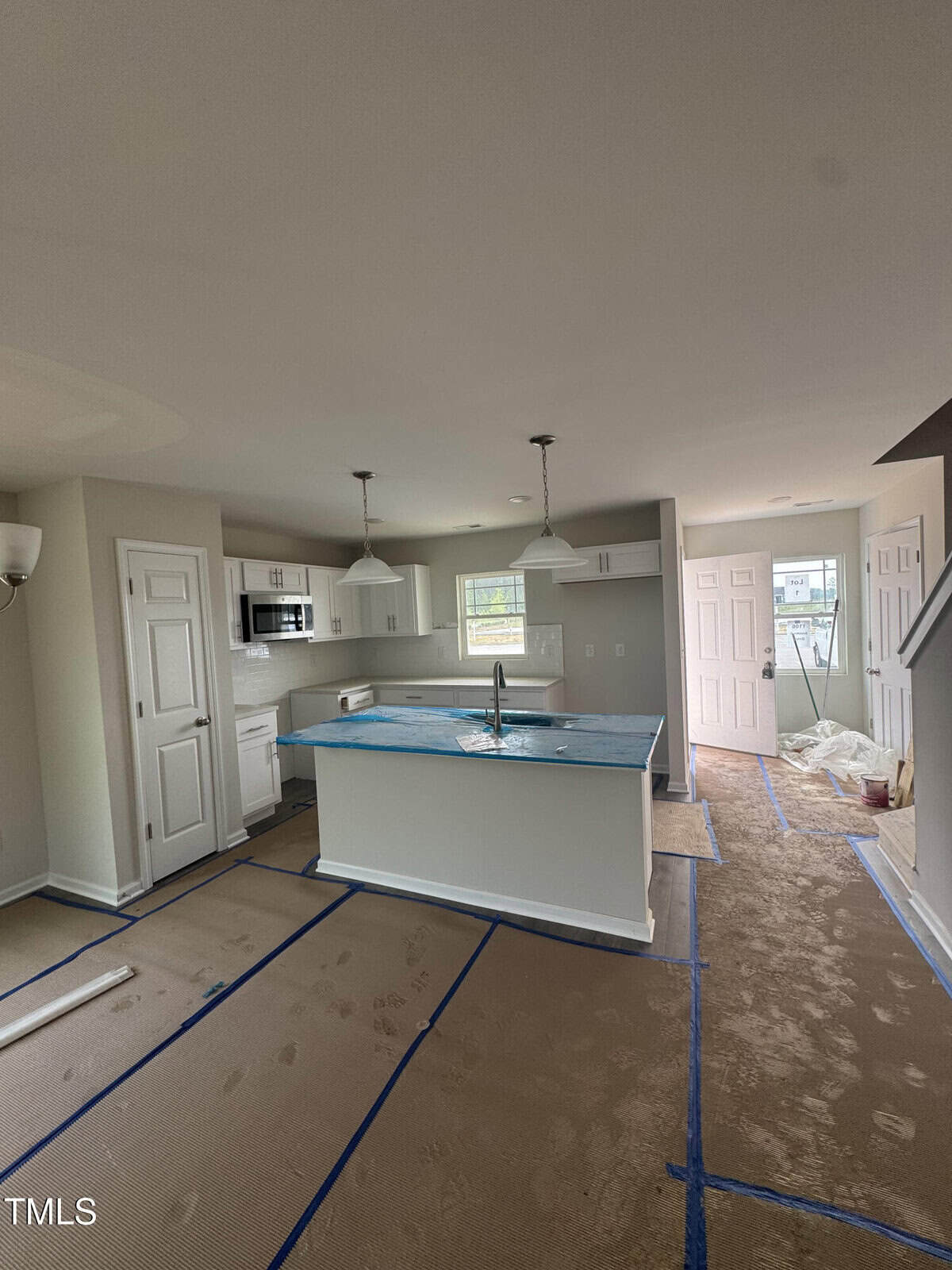 A modern kitchen and living space under construction, featuring an island with a sink, pendant lights, appliances installed, protective coverings on the floors and counters, and construction materials scattered around.
