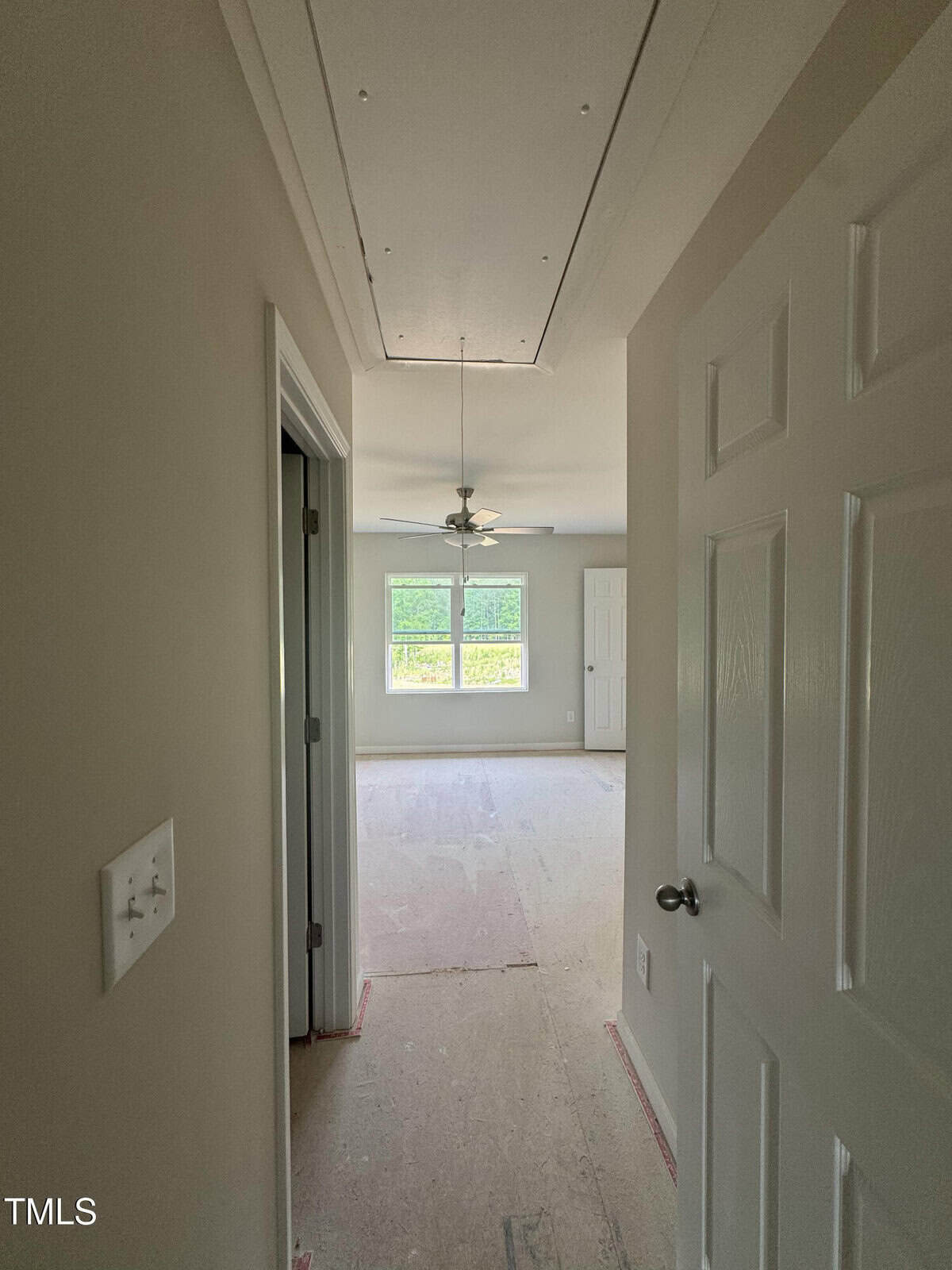 A view from an open doorway into an unfurnished room with a ceiling fan, large window, and bare floor. A hallway leads past a light switch and another open door on the left.