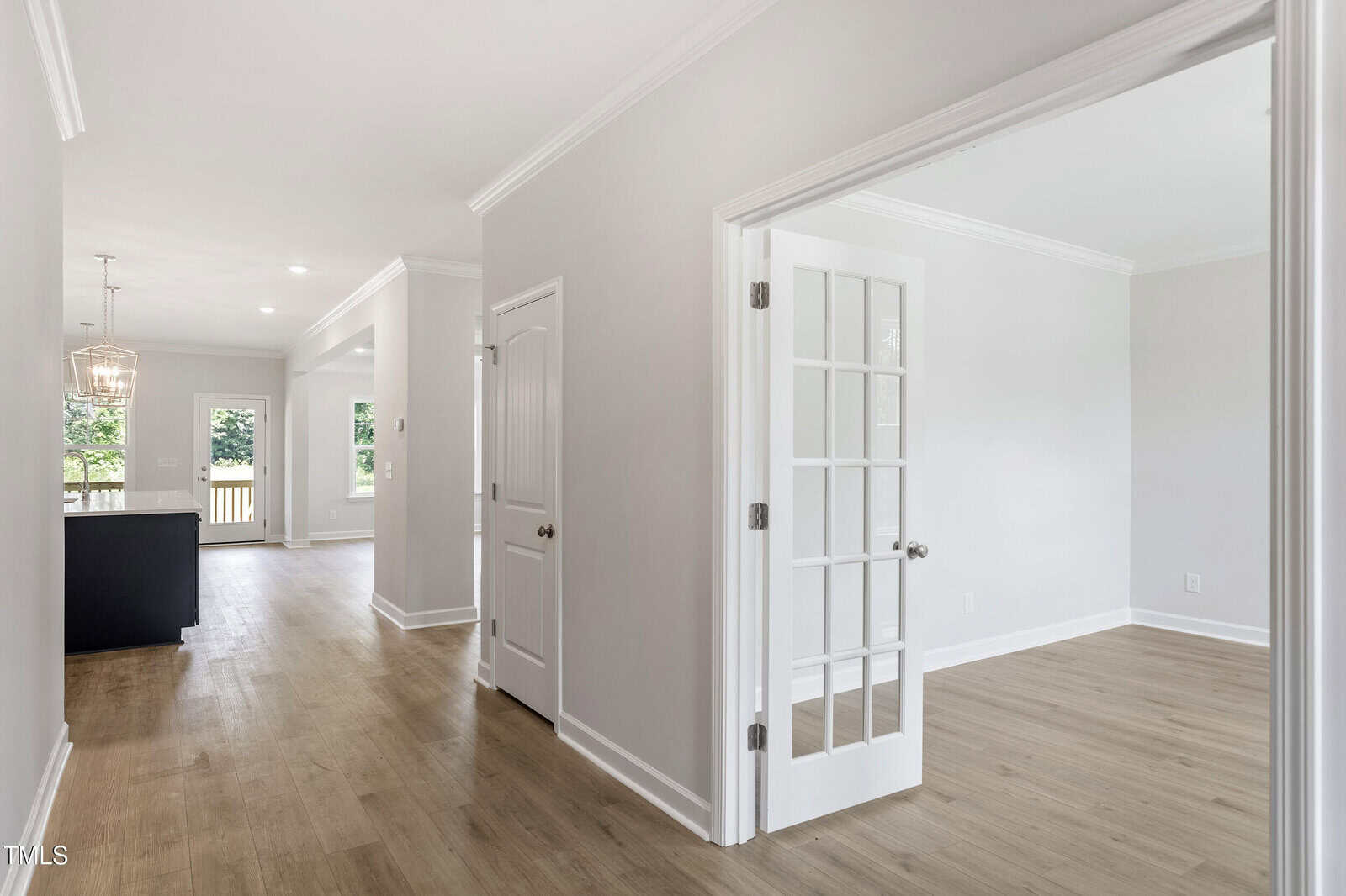 Bright, modern home interior with light wood flooring, white walls, and crown molding. French doors open to a room on the right. Hallway leads to a kitchen and dining area with large windows and pendant lighting.