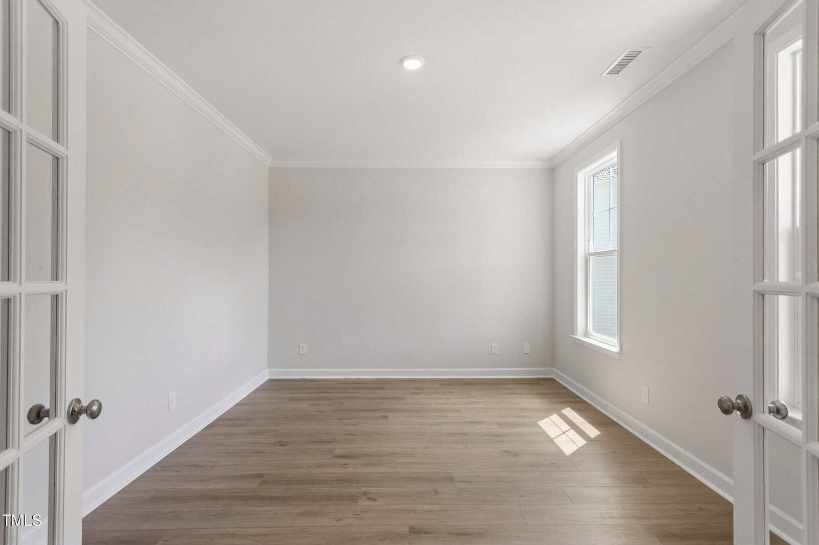 A bright, empty room with light gray walls, wood flooring, and a large window letting in sunlight. French doors with glass panels are partially open in the foreground.