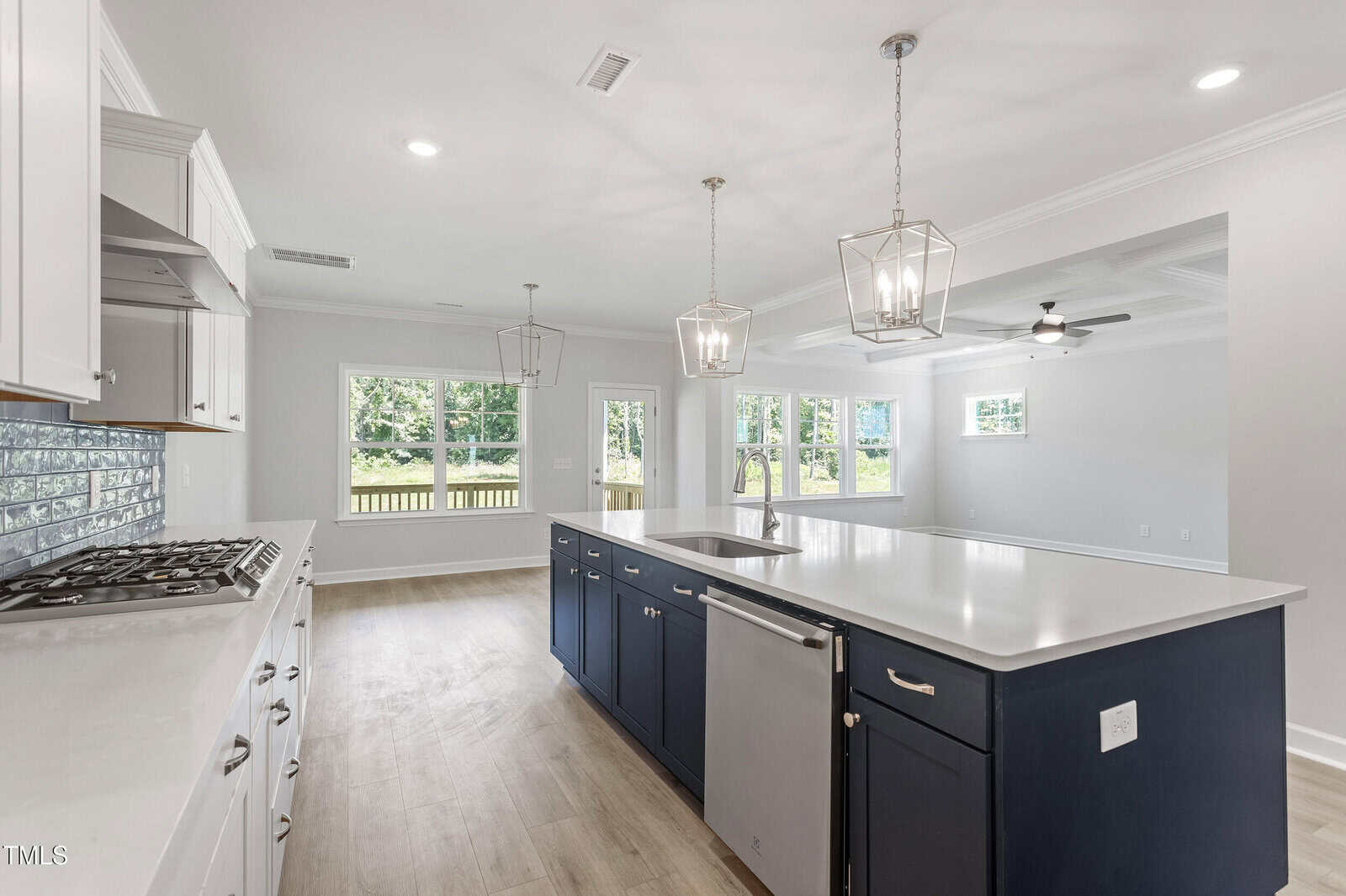 Bright, modern kitchen with a large navy island, white countertops, stainless steel appliances, pendant lights, and open layout leading to a living area with large windows and views of greenery outside.