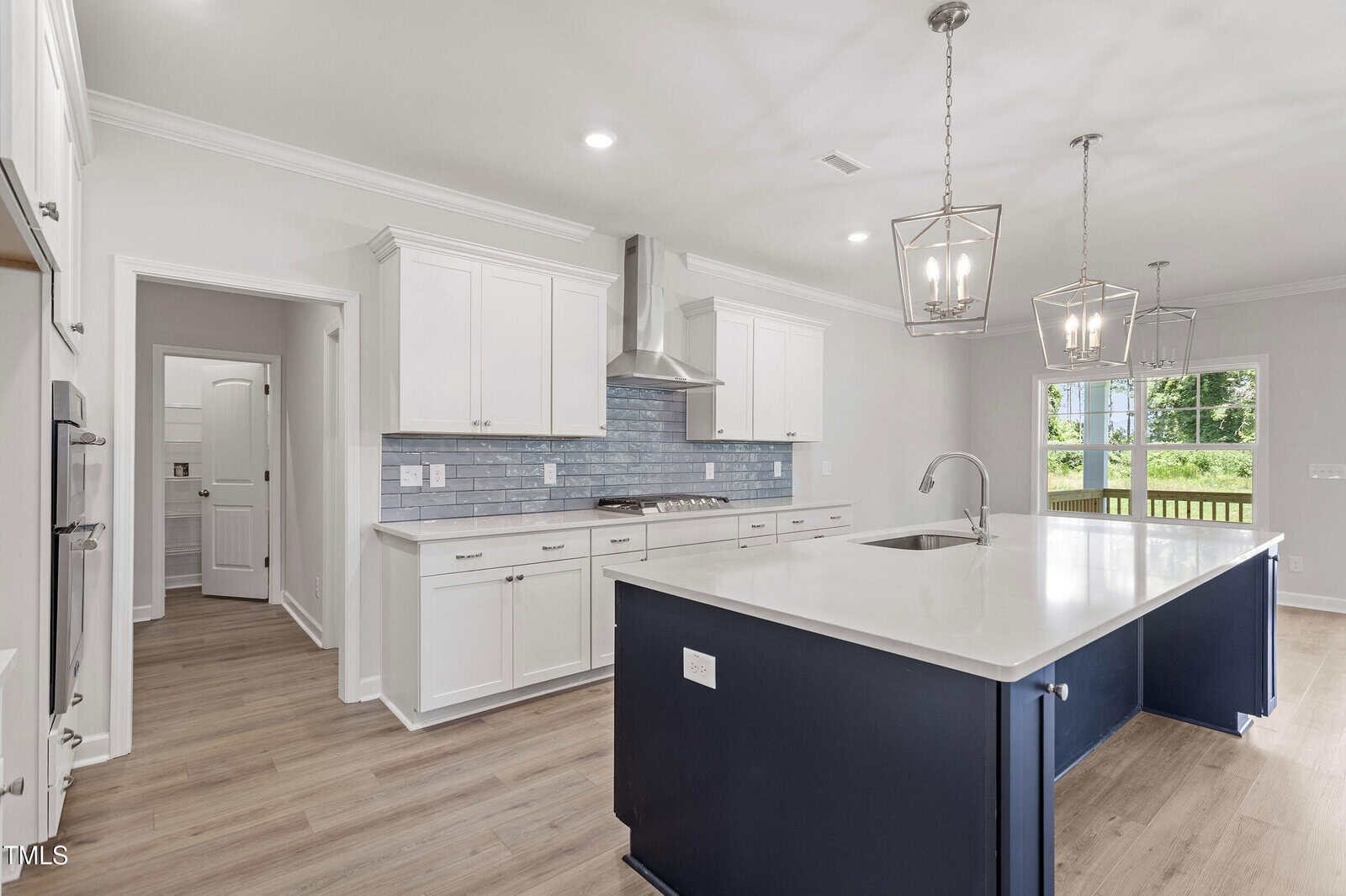 Modern kitchen with white cabinetry, a blue island with a sink, stainless steel appliances, light wood floors, blue tile backsplash, and large windows letting in natural light. Two pendant lights hang above the island.