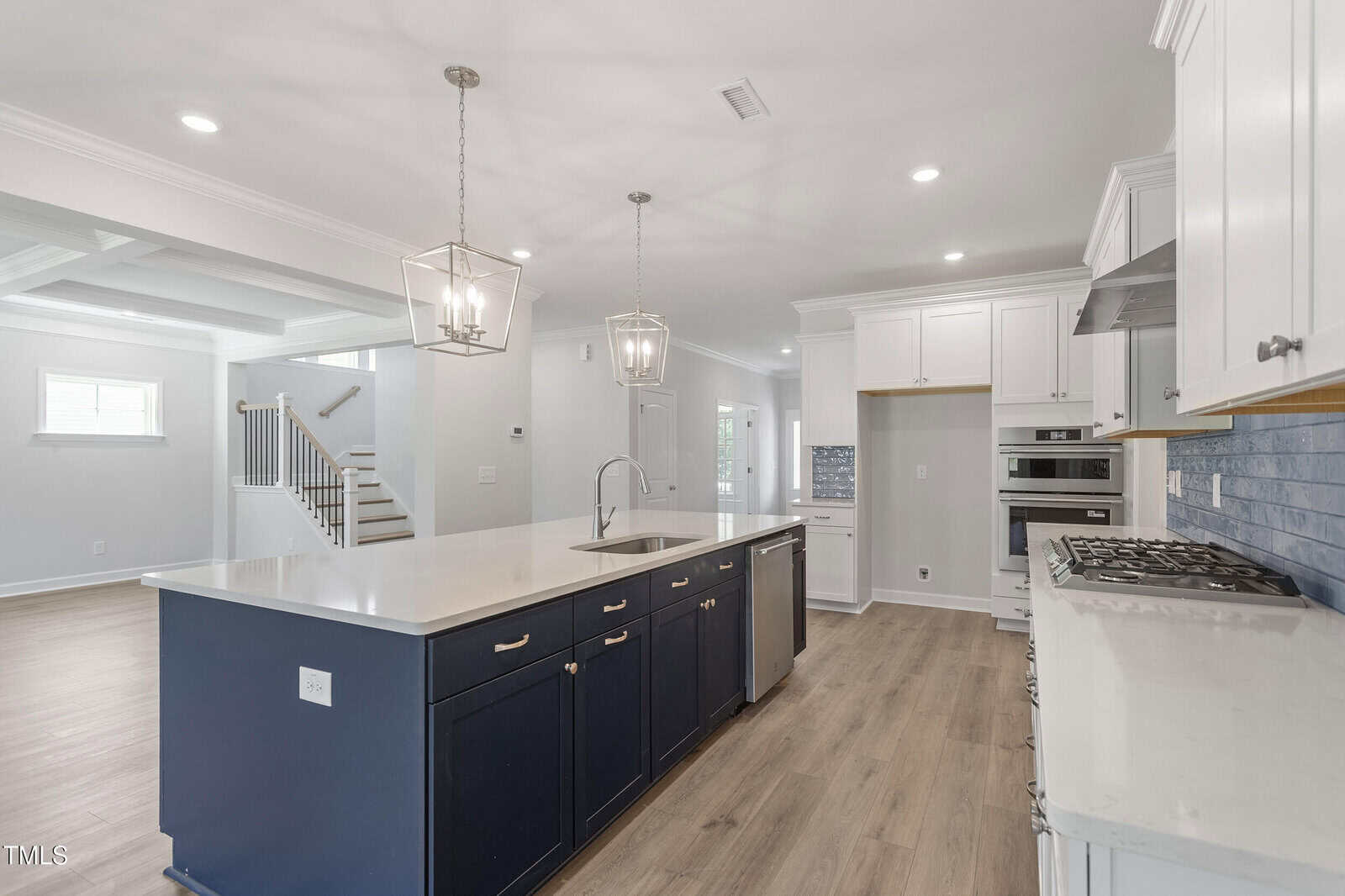 Modern kitchen with navy blue island, white countertops, stainless steel appliances, white cabinetry, wood flooring, pendant lights, and an open layout connecting to a bright living area and stairway.