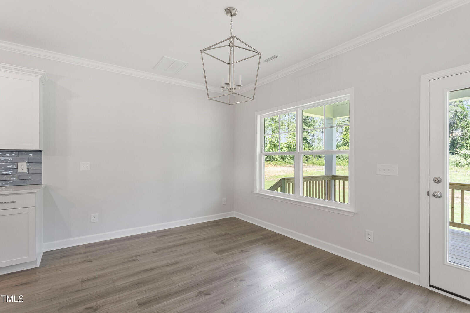 A bright, empty room with light wood flooring, light gray walls, a large window, a modern geometric pendant light, and a glass door leading to a porch with green grass and trees outside.