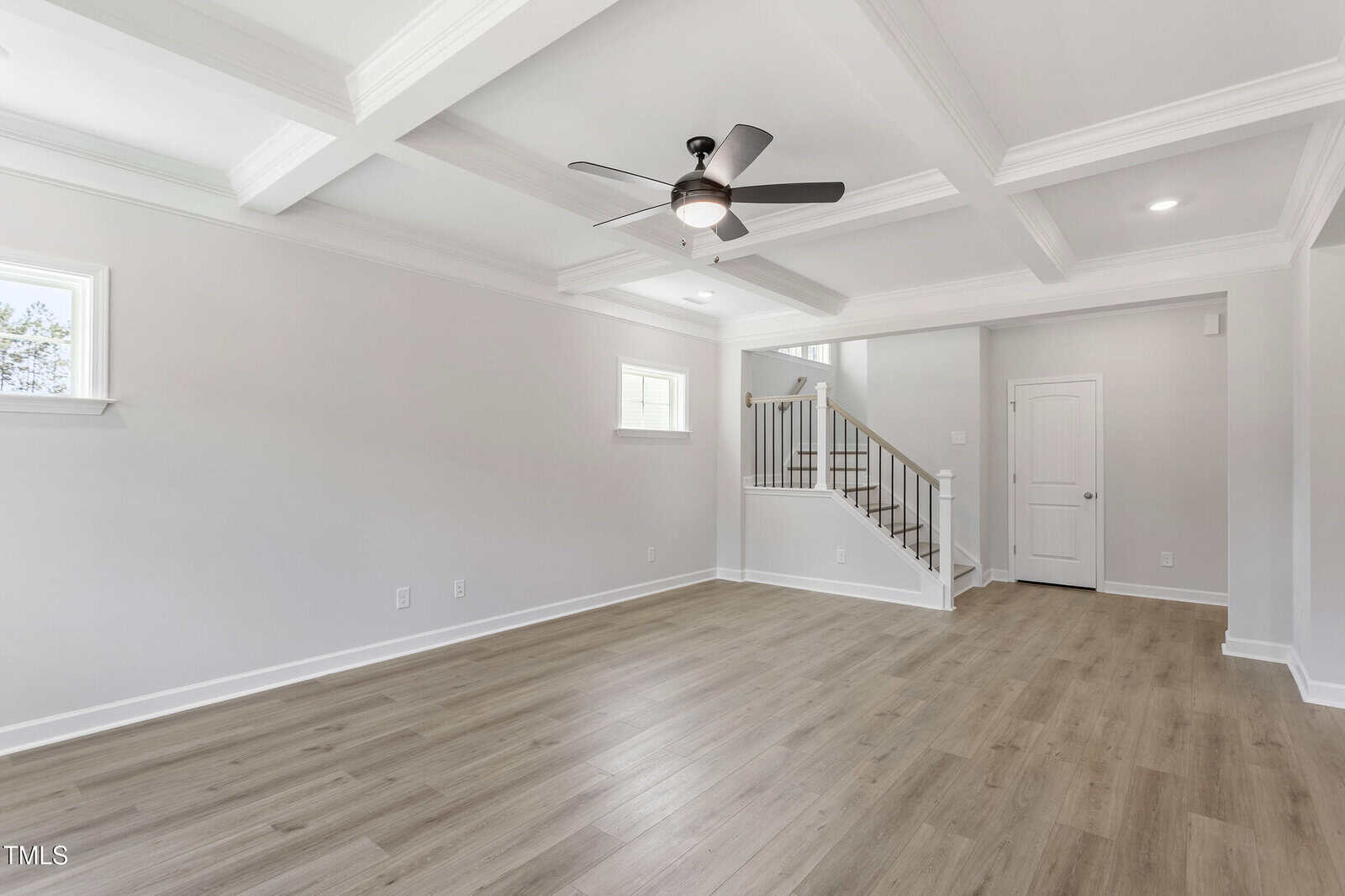 A bright, empty living room with light wood flooring, white walls, coffered ceiling, a ceiling fan, recessed lighting, and a staircase with a wooden handrail leading upstairs.