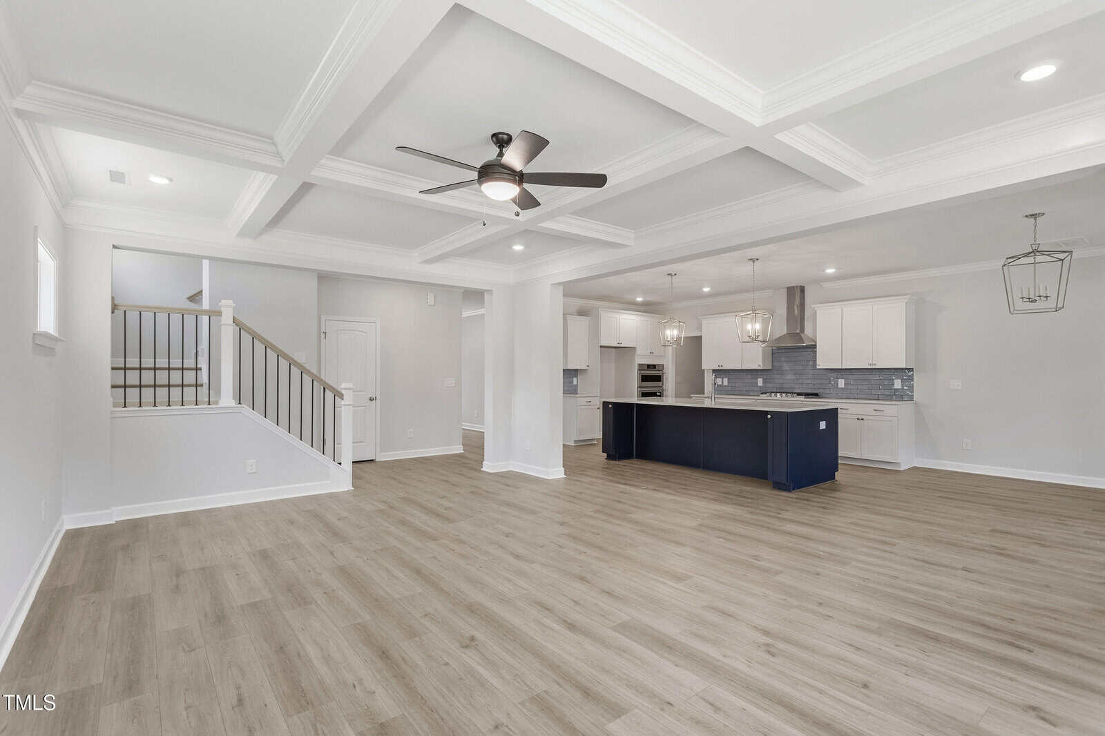 Open-concept living area with light wood flooring, white walls, coffered ceiling, ceiling fan, and recessed lighting. Kitchen features white cabinets, dark island, stainless steel appliances, and pendant lights. Stairs on the left.