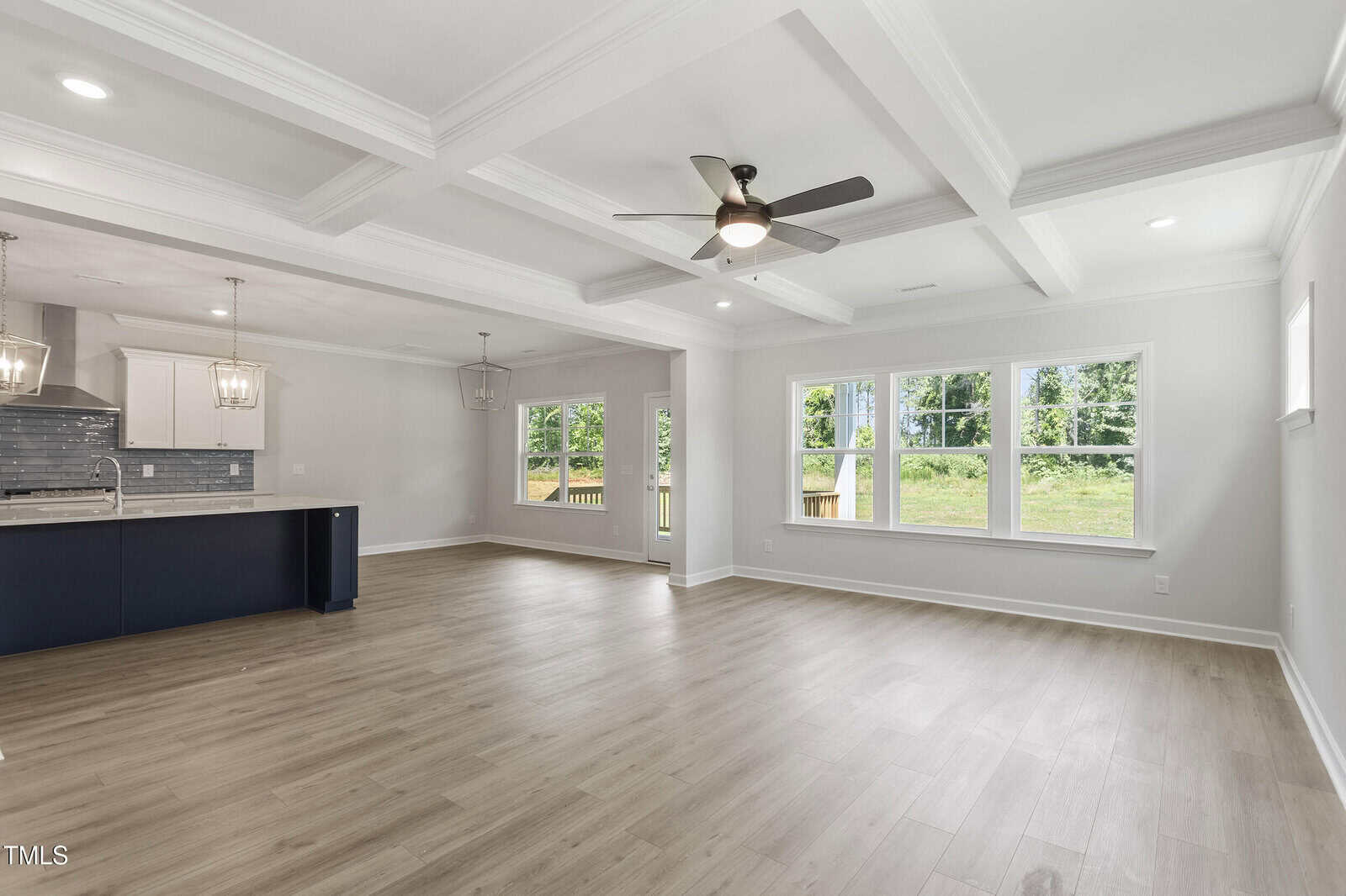Open-concept living area with light wood flooring, coffered ceiling, ceiling fan, large windows, and a modern kitchen featuring a navy island and pendant lights. Outdoors and greenery are visible through the windows.