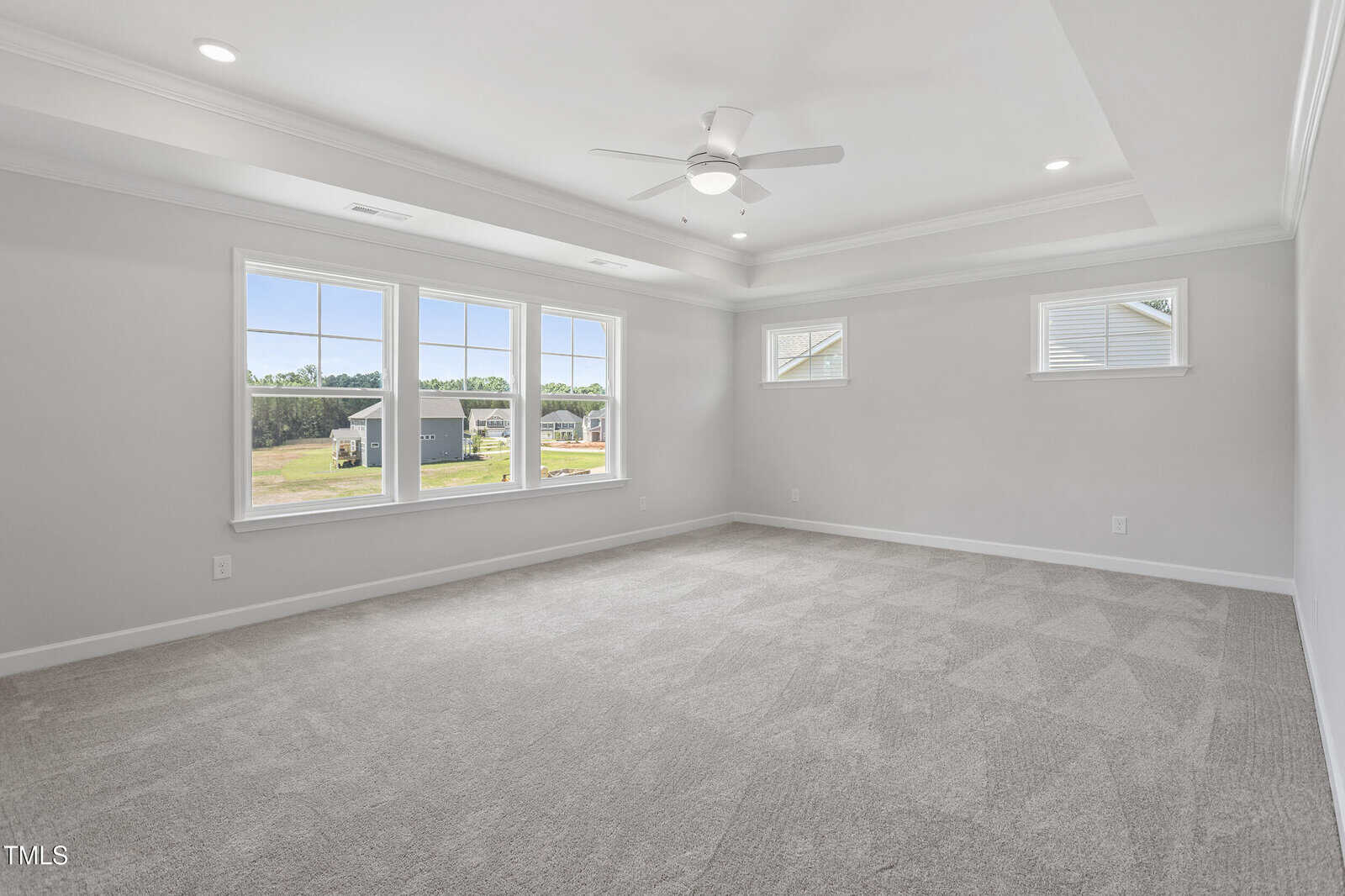 Spacious, empty room with light gray carpet, white walls, tray ceiling, ceiling fan, and large windows letting in natural light. Houses and greenery are visible outside the windows.