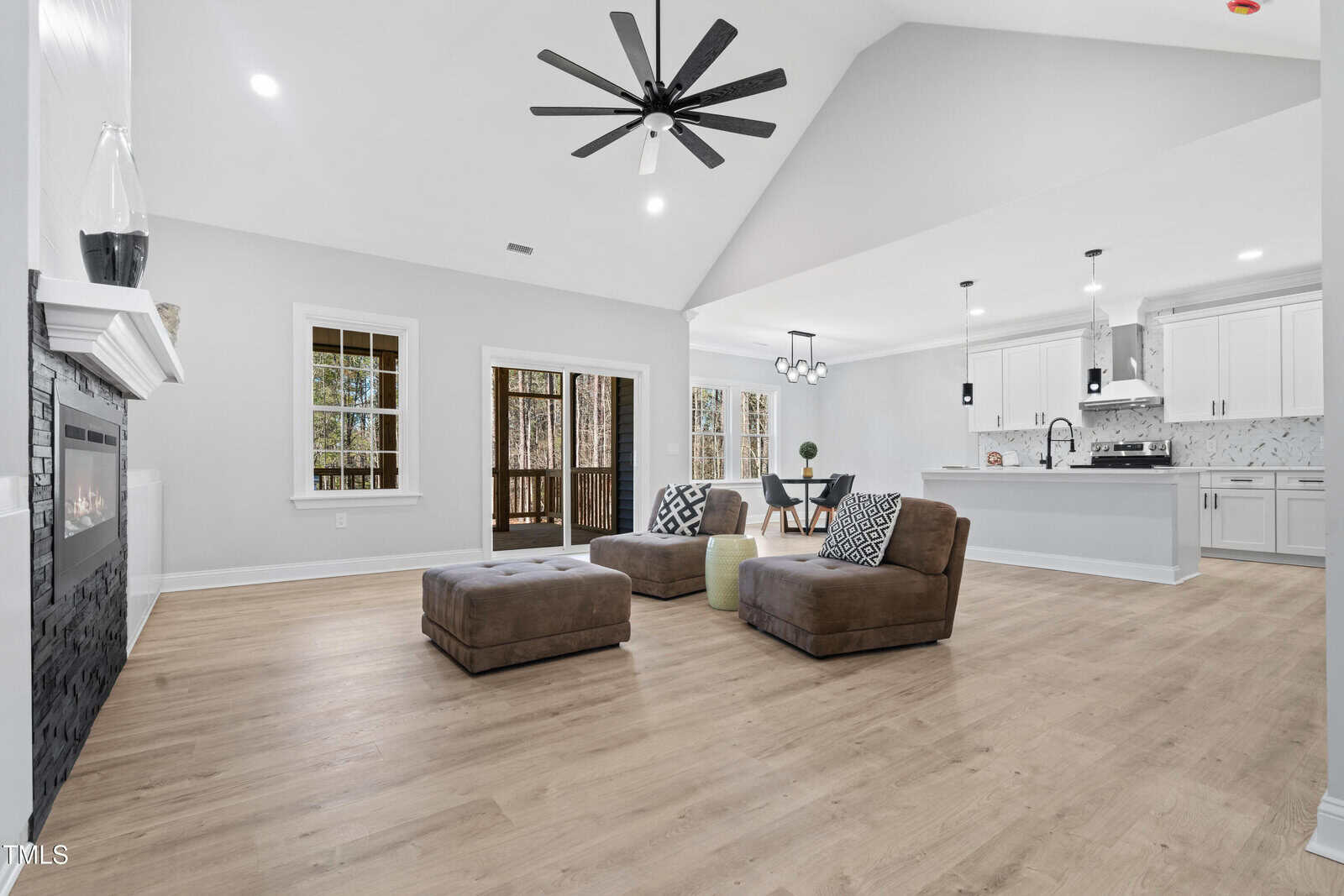 Spacious open-concept living area with wood flooring, two brown ottomans, a fireplace, and ceiling fan. Kitchen with white cabinets and dining area visible. Large windows and doors open to a covered porch.
