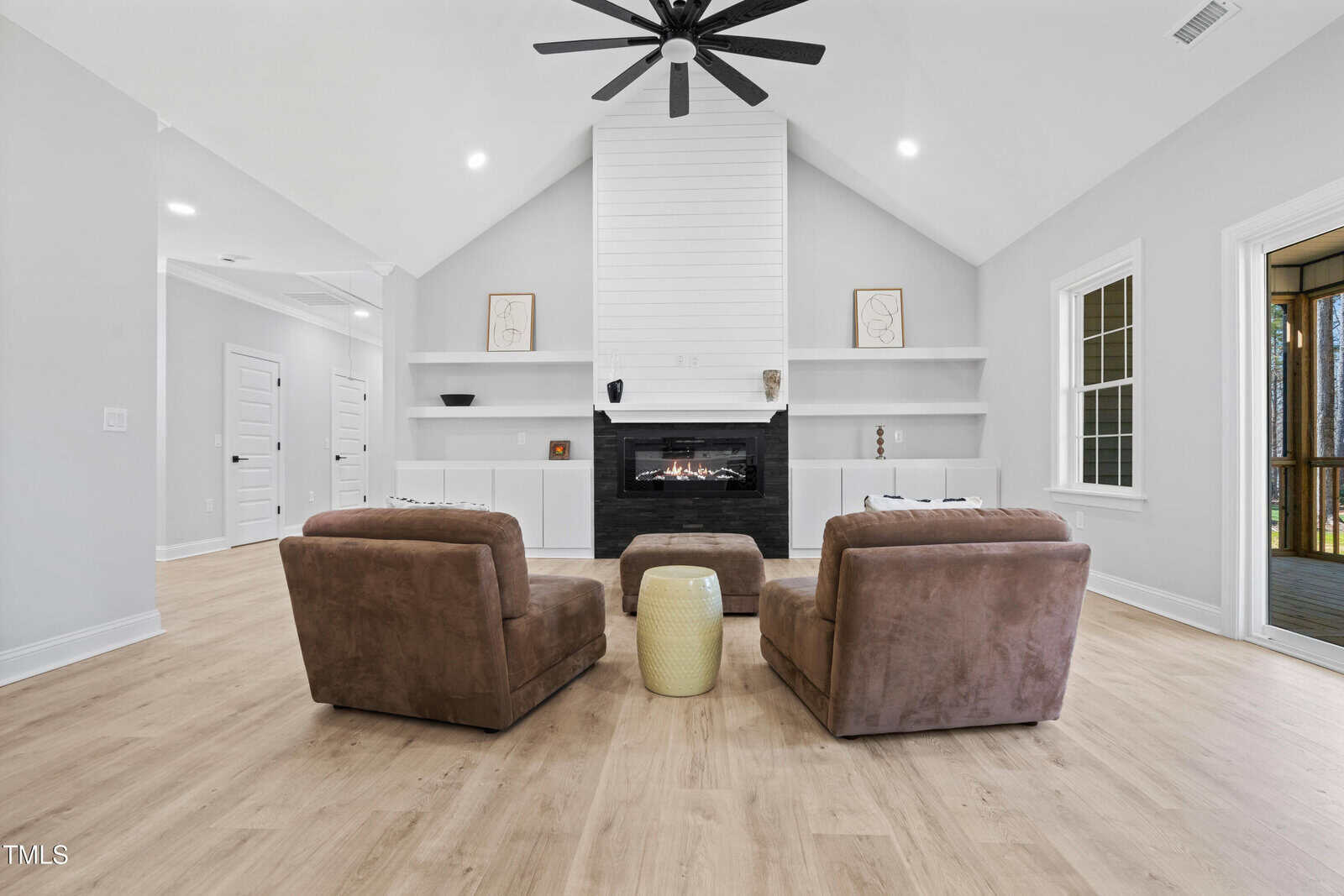 Modern living room with light wood floors, two brown armchairs, a matching ottoman, a small round yellow table, a black fireplace, and a high white vaulted ceiling with a black ceiling fan.