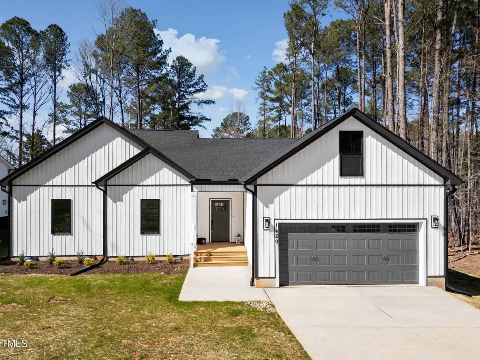 Charming single-story house located at 1429 Sagamore Drive in Louisburg, featuring white vertical siding, black trim, and a dark gray roof. There is a two-car garage, a small front porch, and a concrete driveway. The grassy yard is accented with young shrubs, while tall pine trees surround the property, enhancing its peaceful setting.