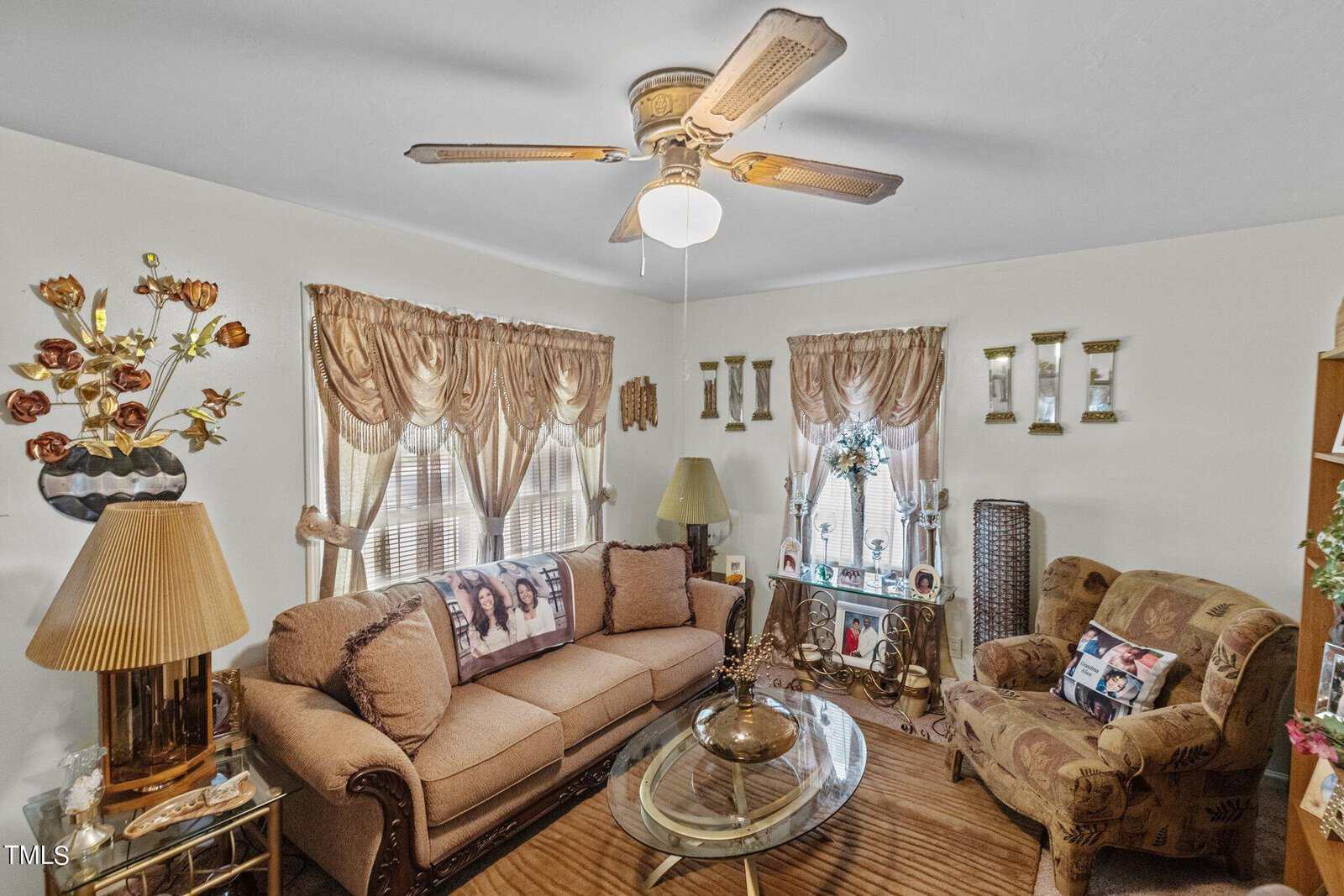 A living room with a ceiling fan and beige walls. It has a sofa with decorative pillows, an armchair, a glass coffee table, and side tables with lamps. Windows feature draped curtains, and various decorative items adorn shelves and tables.