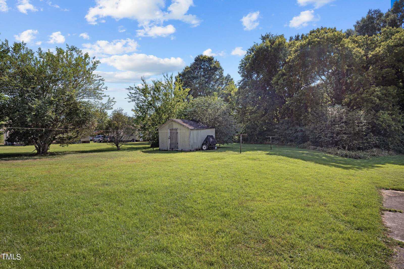 A grassy backyard with a small wooden shed near the center. Trees and bushes are in the background, and a clothesline runs horizontally across the yard. The sky is clear with a few clouds.