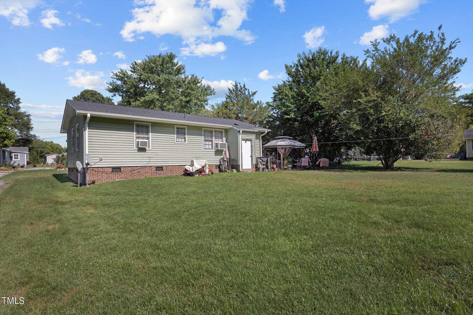 A green single-story house with a sloped roof, brick foundation, and white trim is situated in a grassy yard. Outdoor furniture, including a table with an umbrella, chairs, and a grill, is set up on a patio next to the house. Trees are in the background.