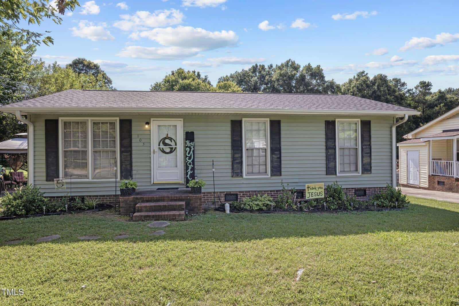 Single-story house with light green siding and dark shutters. Three front windows and a door have decorative elements. Small front garden with a "Thank you, Jesus" sign. Grassy lawn and trees in the background under a blue sky with clouds.