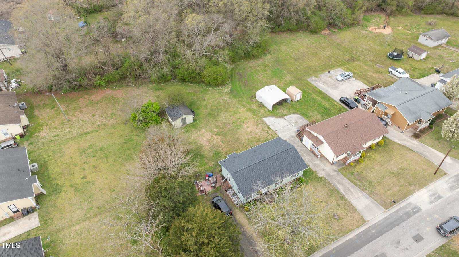 Aerial view of a residential area featuring several houses with yards. One house has a light blue-gray roof and a patio. Nearby, there is a small shed and a canopy with vehicles parked on a driveway. Surrounding trees and grass are visible.