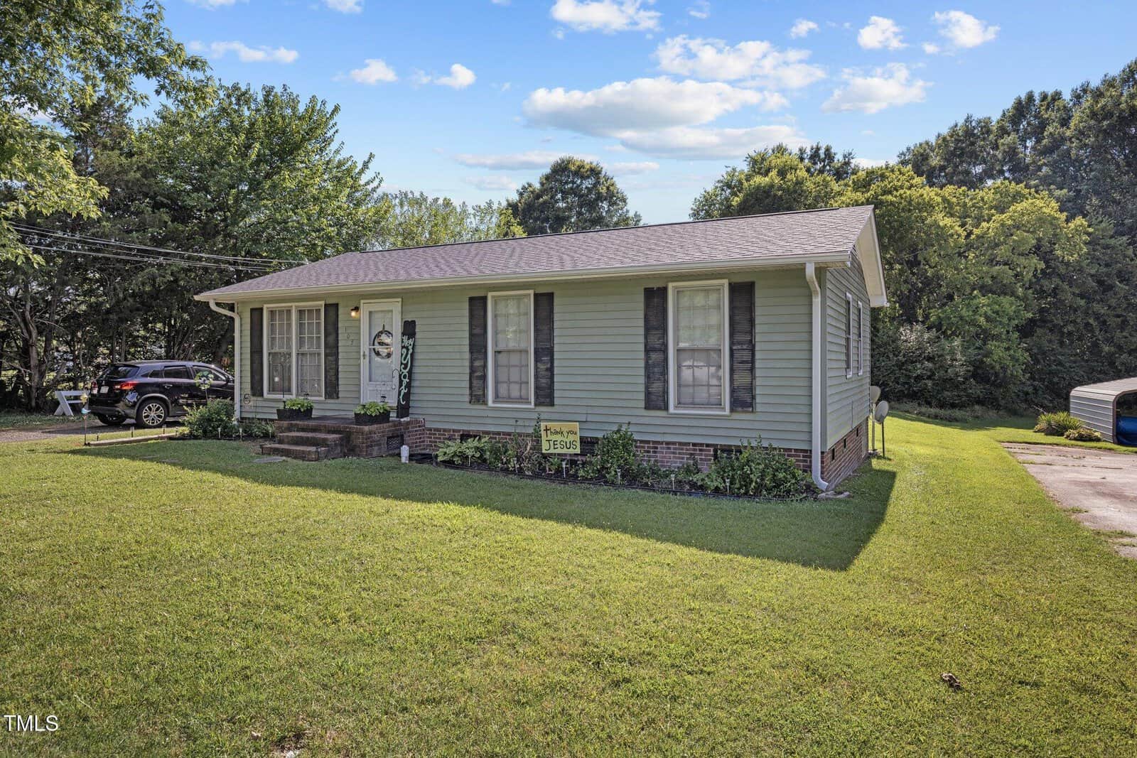 A single-story, light green house with a dark roof, surrounded by a well-maintained lawn. A driveway on the right leads to a parked car and a small outbuilding. Bushes line the front, and trees are in the background under a partly cloudy sky.