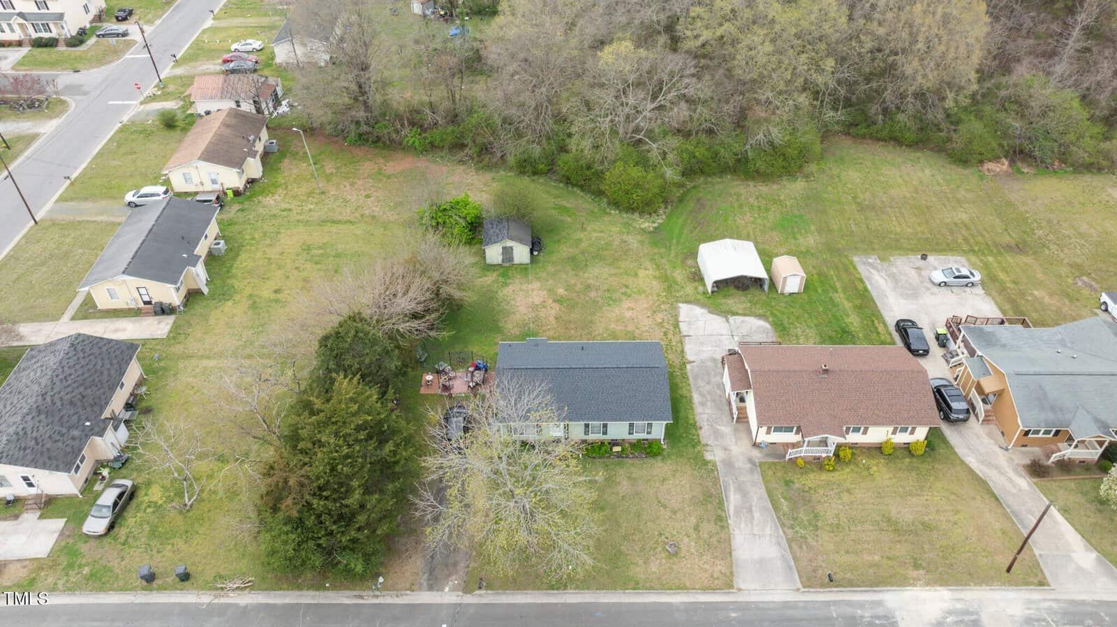 Aerial view of a suburban neighborhood with several houses and a tree-lined area. A group of people is gathered in the backyard of a house near a patio. Driveways are visible, and there is a shed and a carport in the green space.
