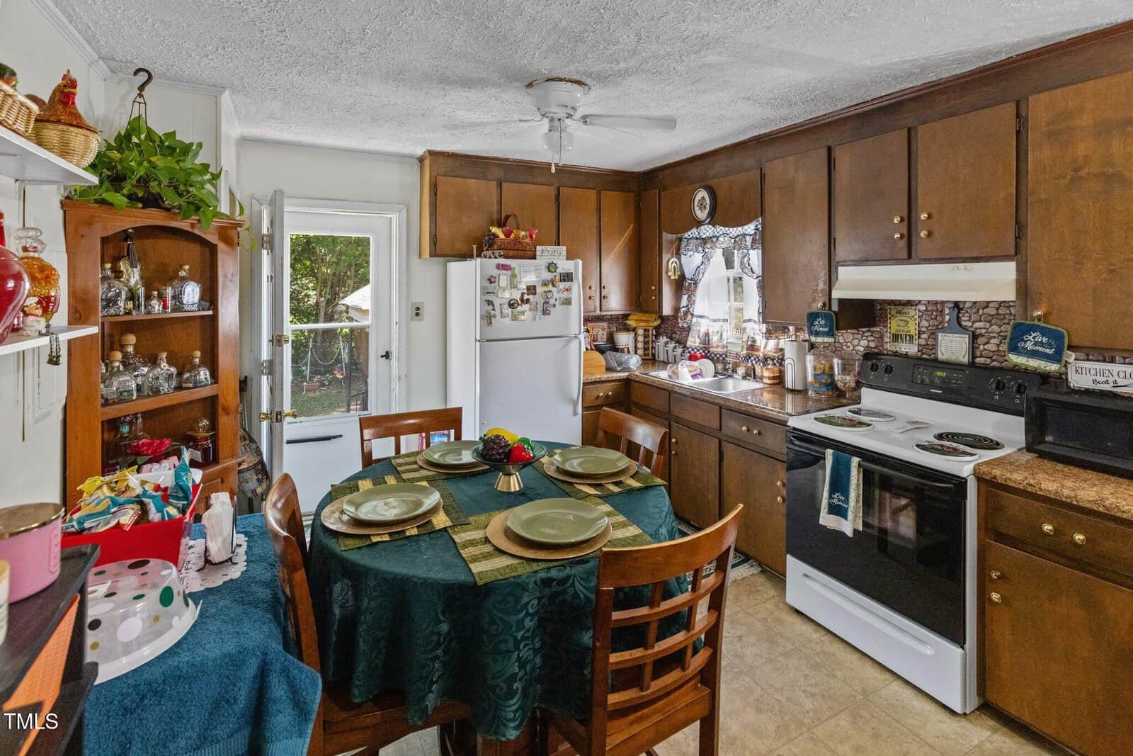 A kitchen with dark wooden cabinets and a dining area. There's a table set for four with green placemats, and a refrigerator with magnets. A stove with a hood is next to a small window. Shelves display various knick-knacks and plants.