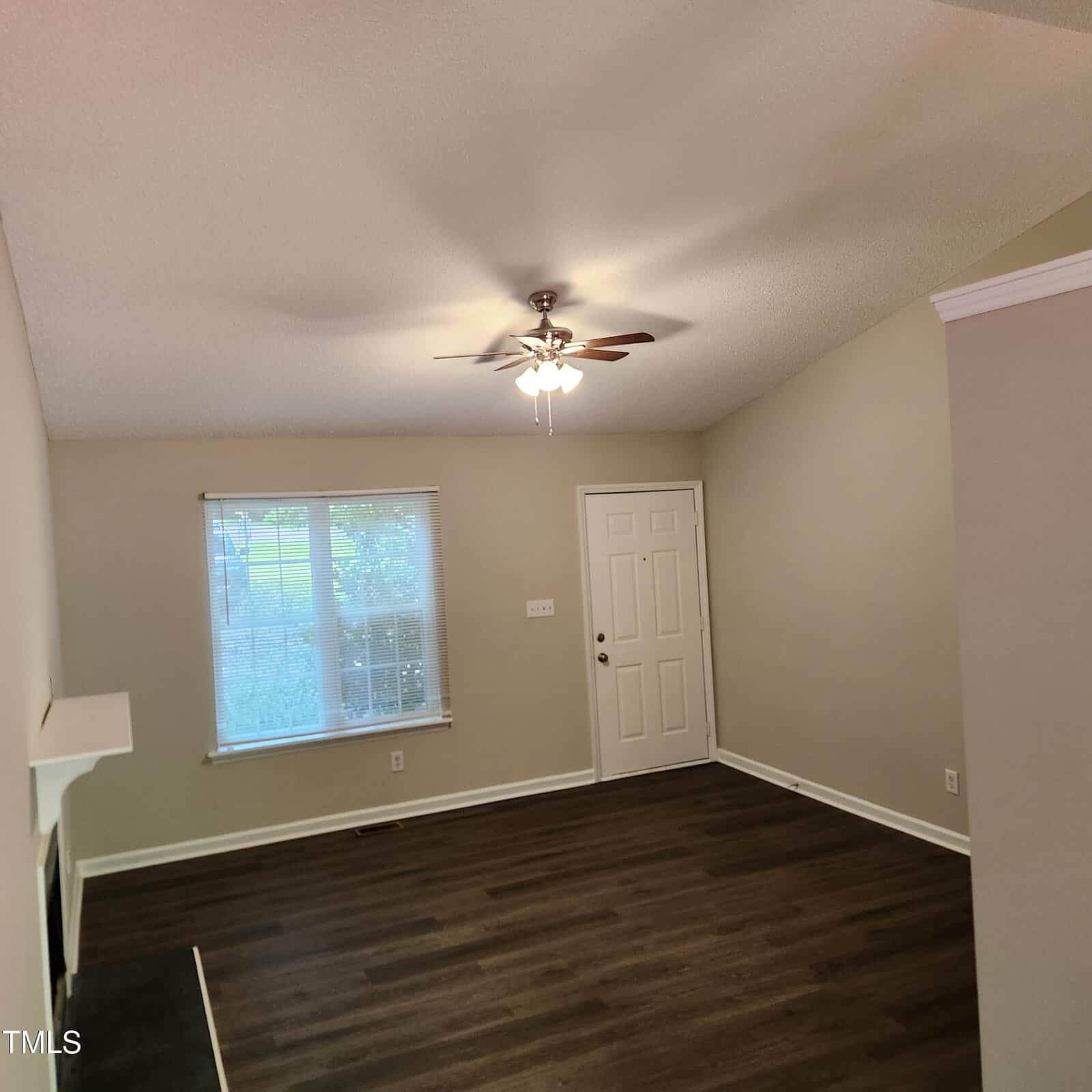 A bright, empty room with light beige walls, a ceiling fan with lights, a window with blinds, a white door, and dark wood flooring. Sunlight is coming in through the window.