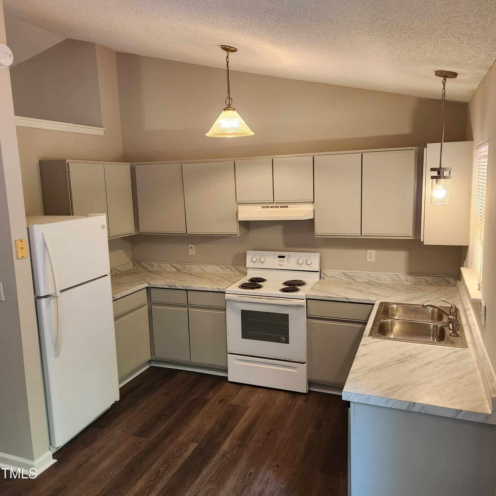 A modern kitchen with light gray cabinets, white appliances including a refrigerator and stove, marble-patterned countertops, double sink, wood-style flooring, and two hanging pendant lights.