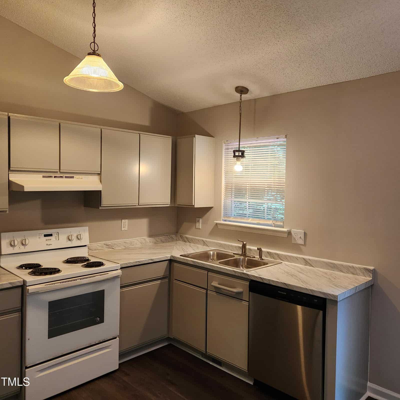 Modern kitchen with beige walls and cabinets, white electric stove, stainless steel dishwasher, double sink under a window with blinds, and two hanging pendant lights. Marble-patterned countertops and dark wood flooring.
