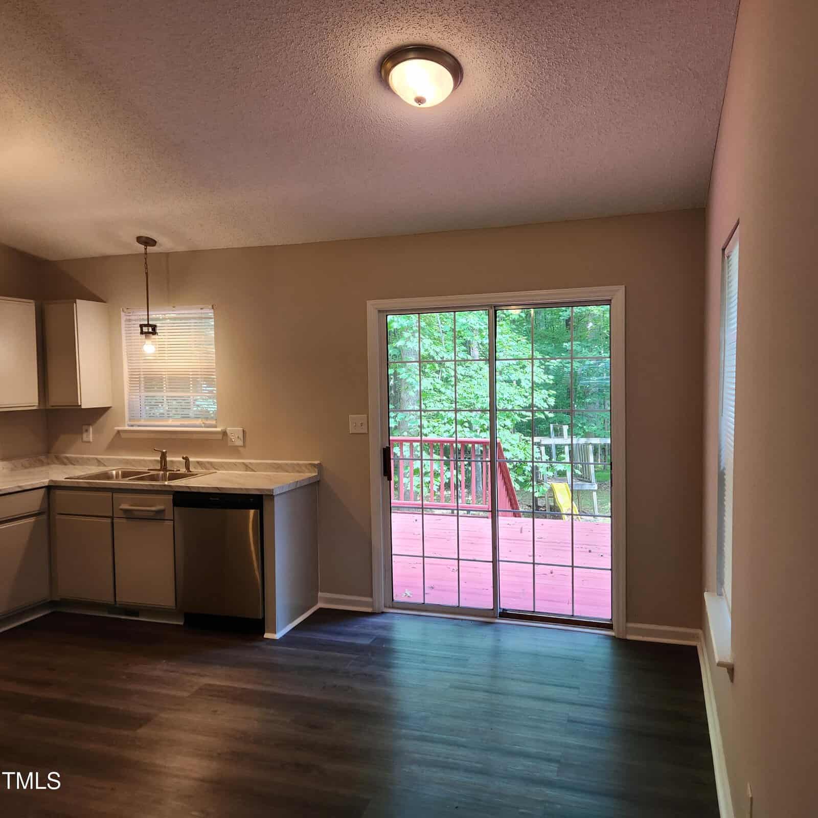 A bright kitchen with white cabinets and dark floors, featuring a stainless steel dishwasher. A sliding glass door opens to a red wooden deck overlooking green trees. A window above the sink lets in more natural light.