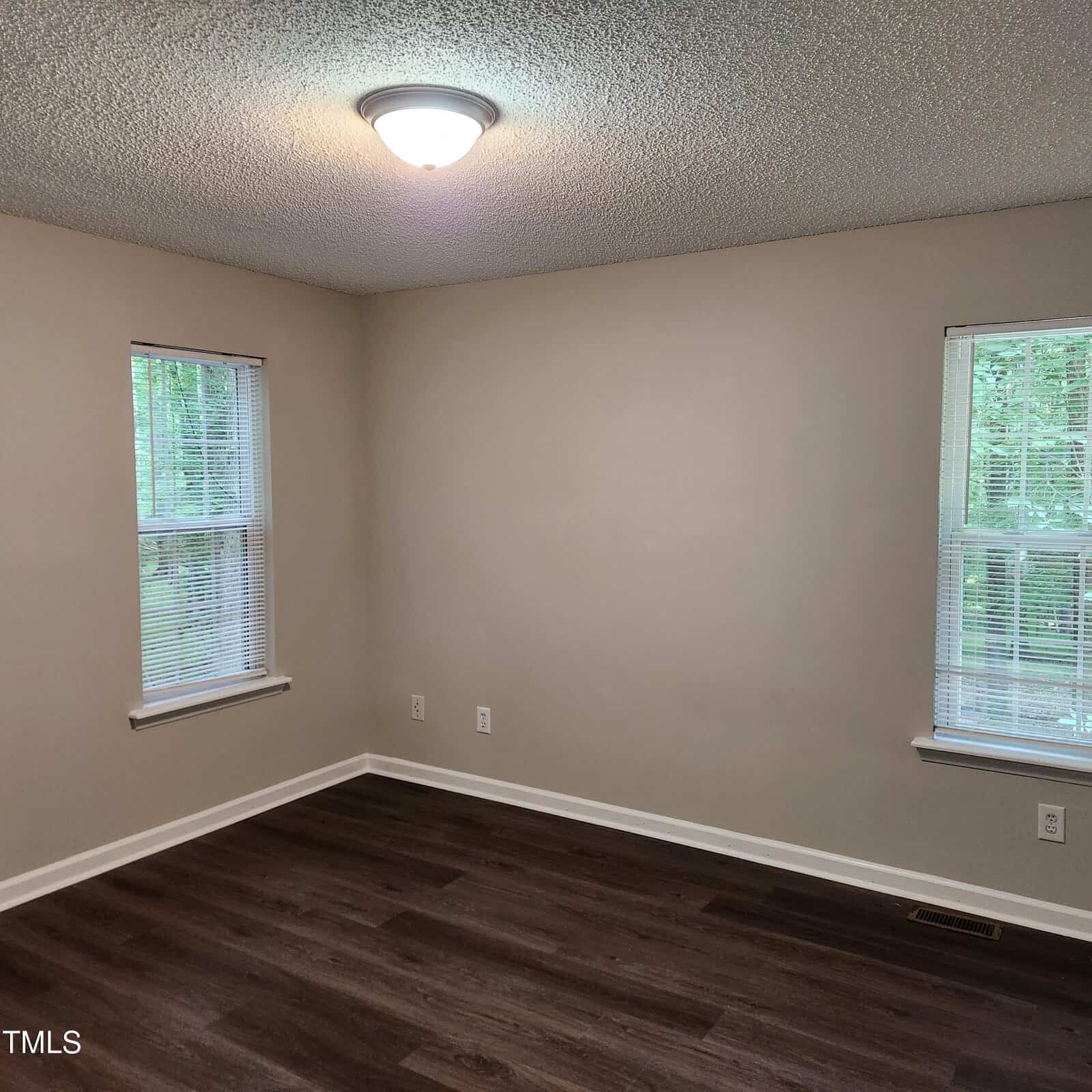Empty room with beige walls, two windows with blinds, dark wood flooring, white baseboards, and a ceiling light. Trees are visible outside the windows.