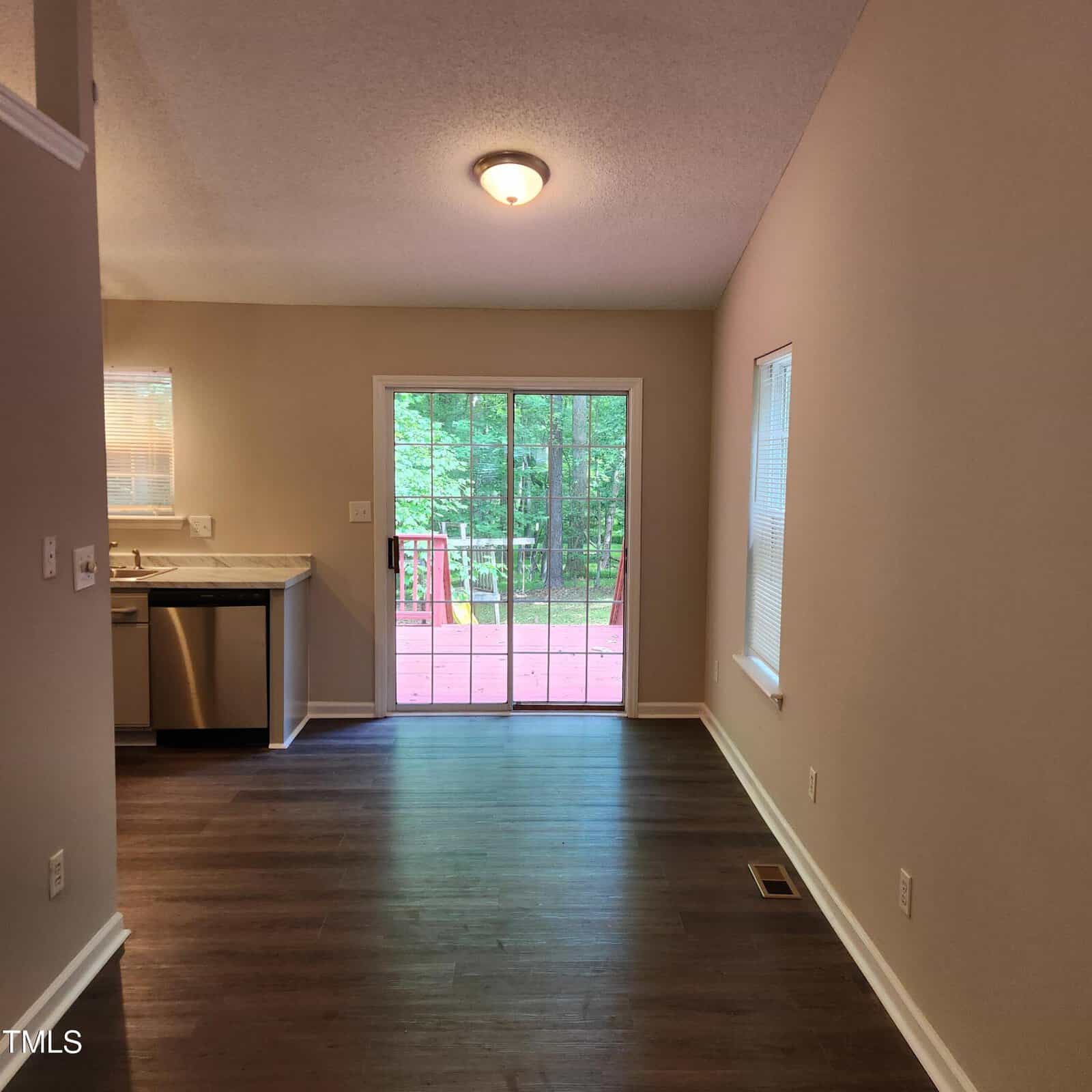 A room with wood flooring, beige walls, and a ceiling light. Sliding glass doors lead to a red deck outside with trees visible. A window is on the right wall, and a kitchen with stainless appliances is on the left.