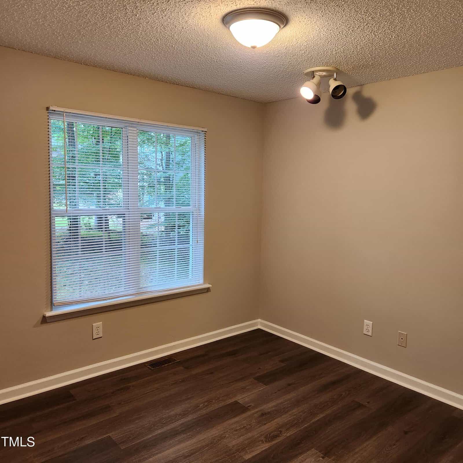 A small empty room with beige walls, a large window with blinds showing trees outside, dark wood flooring, a ceiling light, and a wall-mounted spotlight. Electrical outlets are visible on the walls.