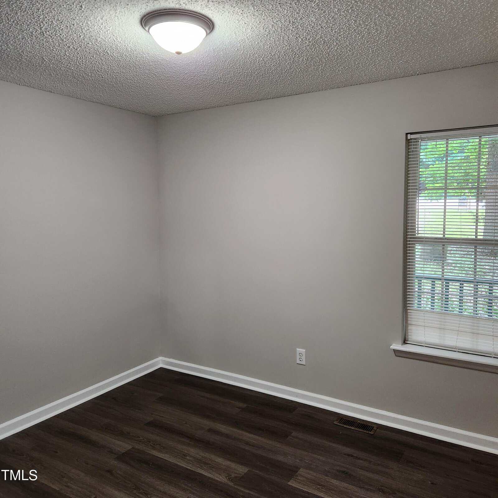 A small, empty room with light gray walls, a white ceiling with a ceiling light, dark wood floor, and a window with blinds letting in natural light. An electrical outlet is on the wall near the window.