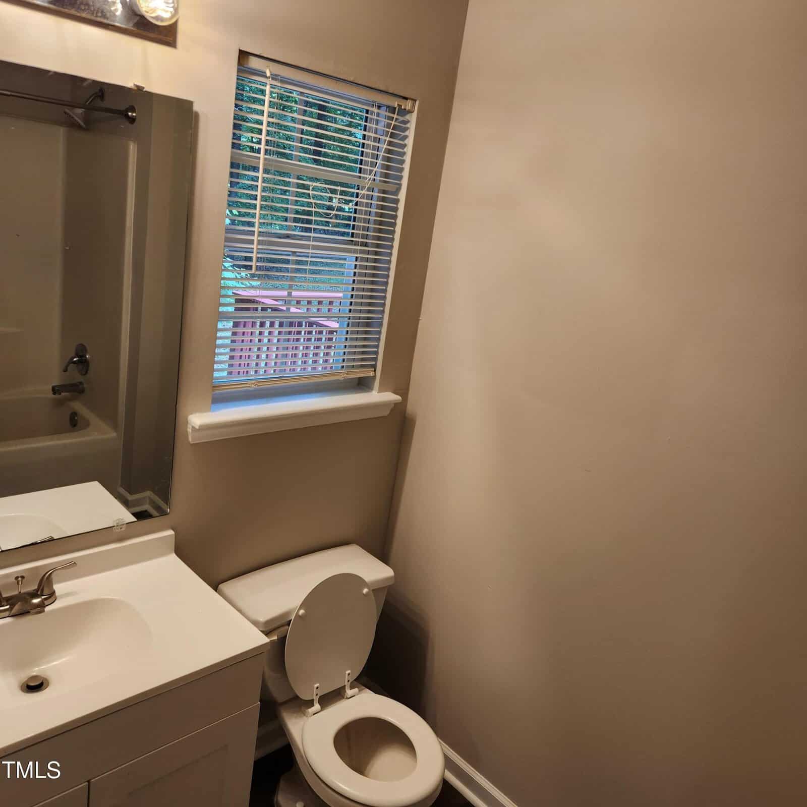 Small bathroom with beige walls, a white vanity and sink, a toilet with the lid open, a window with blinds, and a mirror reflecting part of a bathtub and shower.
