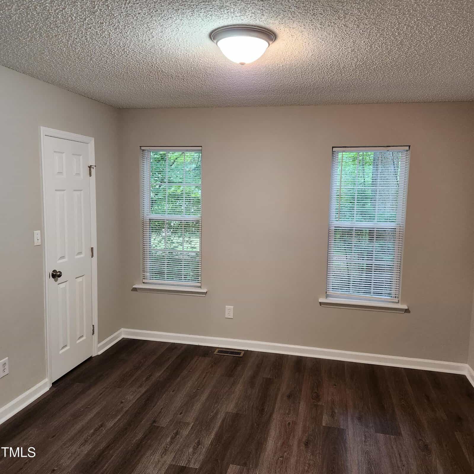 Empty room with beige walls, two windows with blinds, a white door, and dark wood flooring. A ceiling light fixture is turned on, and trees are visible outside through the windows.