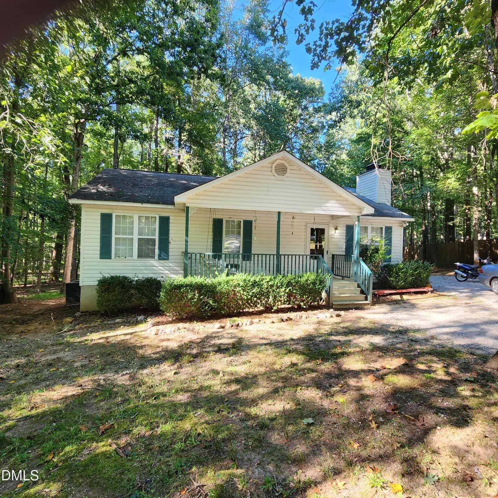 A single-story house with white siding, green shutters, a front porch with railings, and steps leading to the entrance. Surrounded by trees, the house sits on a shaded lot with a gravel driveway.
