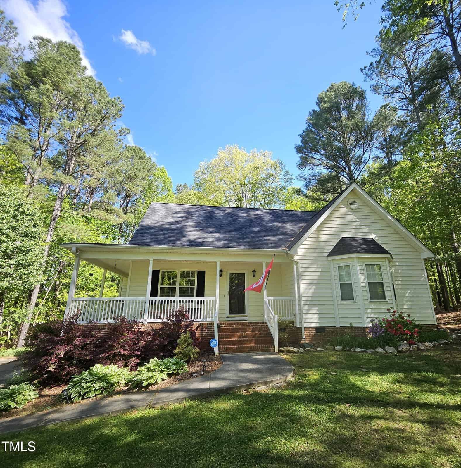 A light yellow single-story house with a front porch, American flag, and gable roof sits on a grassy lot surrounded by tall trees and landscaping on a sunny day.