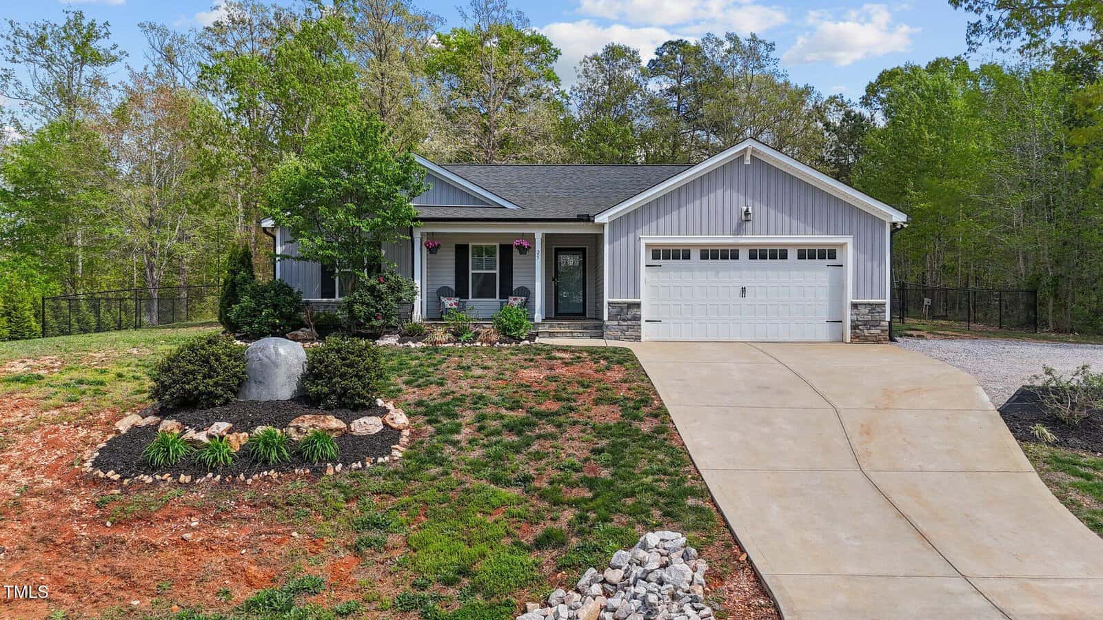 A single-story house with gray siding and a white garage door, a covered front porch with columns, and a landscaped yard featuring mulch, rocks, and shrubs. A concrete driveway leads to the garage. Trees surround the property.