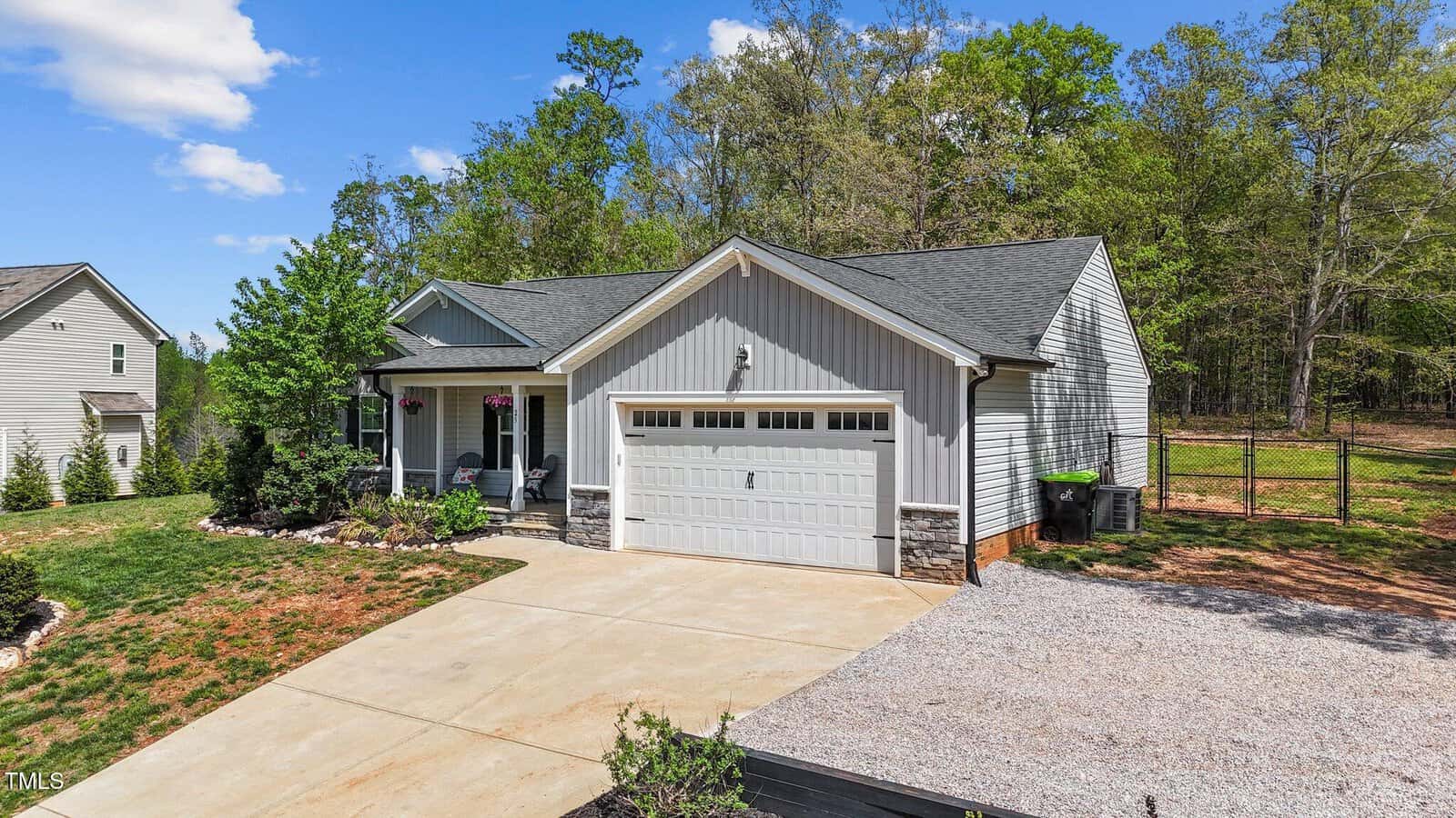 A single-story house with gray siding, a two-car garage, a covered front porch, and a gravel driveway extension. The yard has minimal landscaping and is bordered by trees in the background under a blue sky.