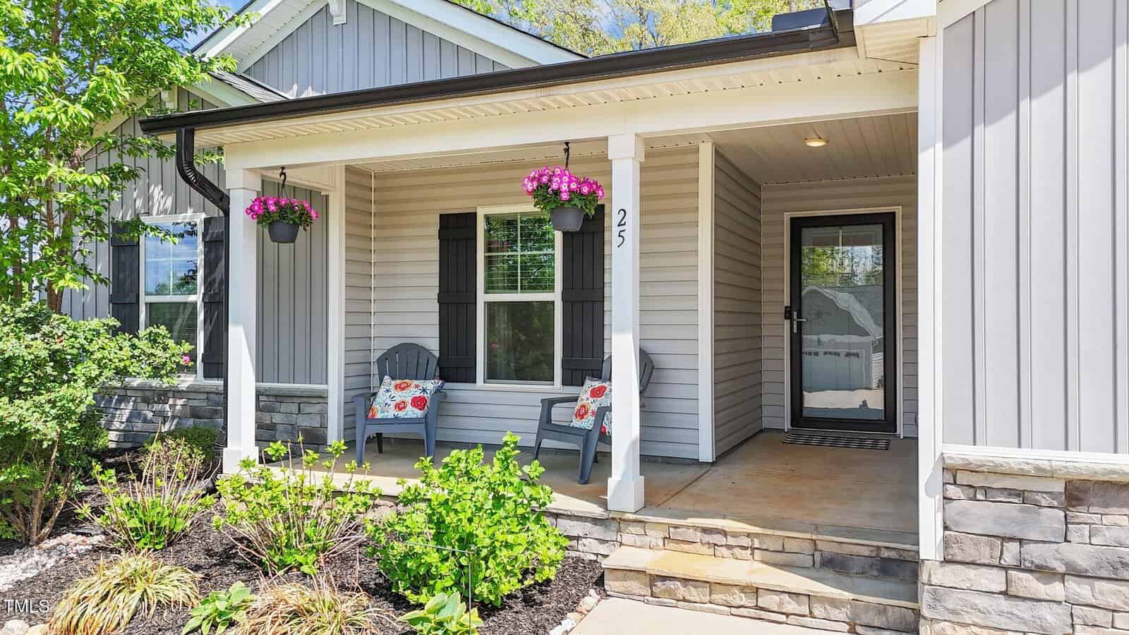 A small front porch with two gray Adirondack chairs, flower pots hanging from the ceiling, a window with black shutters, and a glass-front door. The porch is surrounded by shrubs and flowering plants.